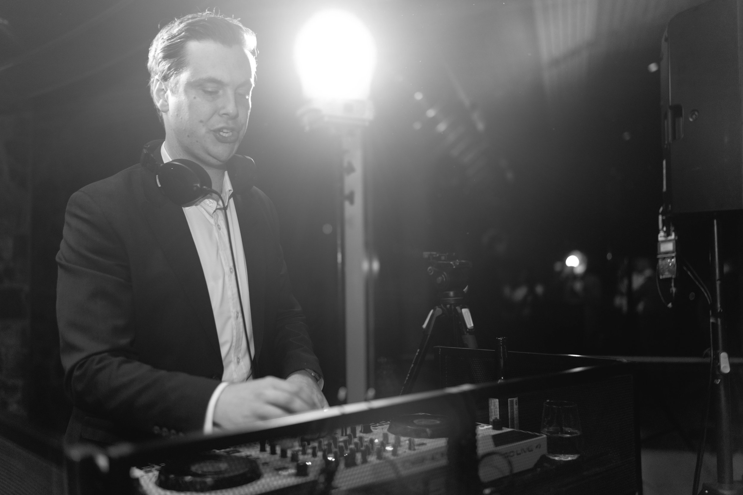 Melbourne DJ in a suit with headphones around his neck, djing wedding, with bright stage lights behind him.