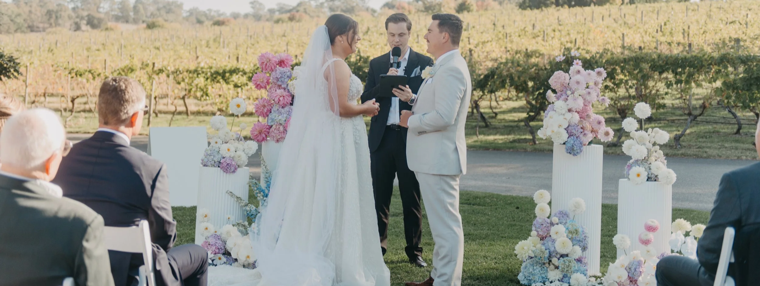 A wedding ceremony taking place outdoors in a vineyard, with a bride and groom exchanging vows in front of an officiant. The bride is in a white wedding dress, and the groom is in a white suit. Floral arrangements with pink, purple, and white flowers decorate the altar. Guests are sitting in chairs, watching the ceremony.