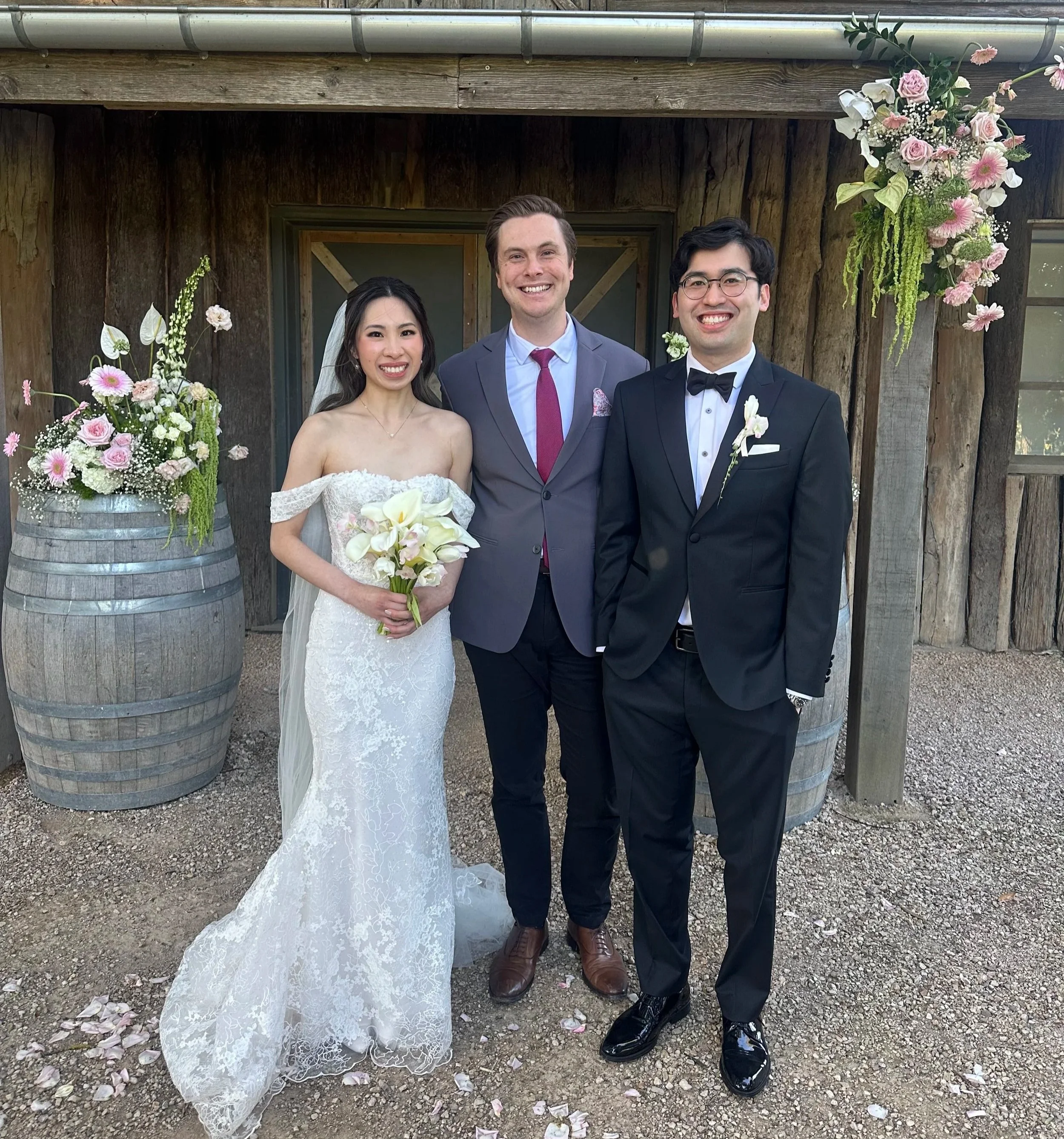 A bride and groom standing with a man in a gray suit at a wedding ceremony, outdoors with wooden background and floral decorations.