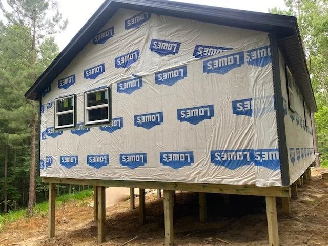 House under construction with Lowes-branded house wrap, elevated on wooden stilts, surrounded by trees.