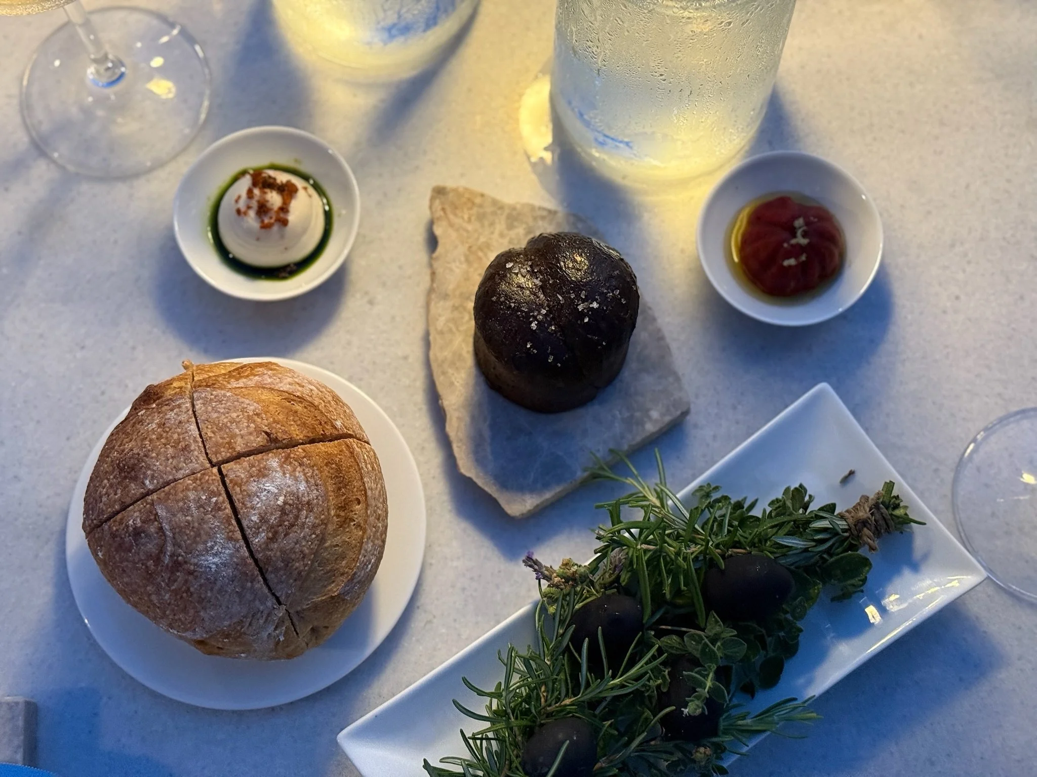 A table with bread, olives, cheese, condiments, a glass of white wine, and a water bottle.