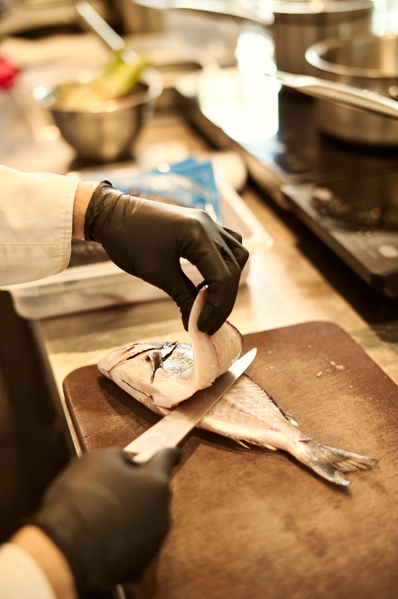 Chef Chris wearing black gloves and a white coat is filleting a fish on a cutting board in a kitchen or market setting. Chefs Table, Agora Restaurant Symi,