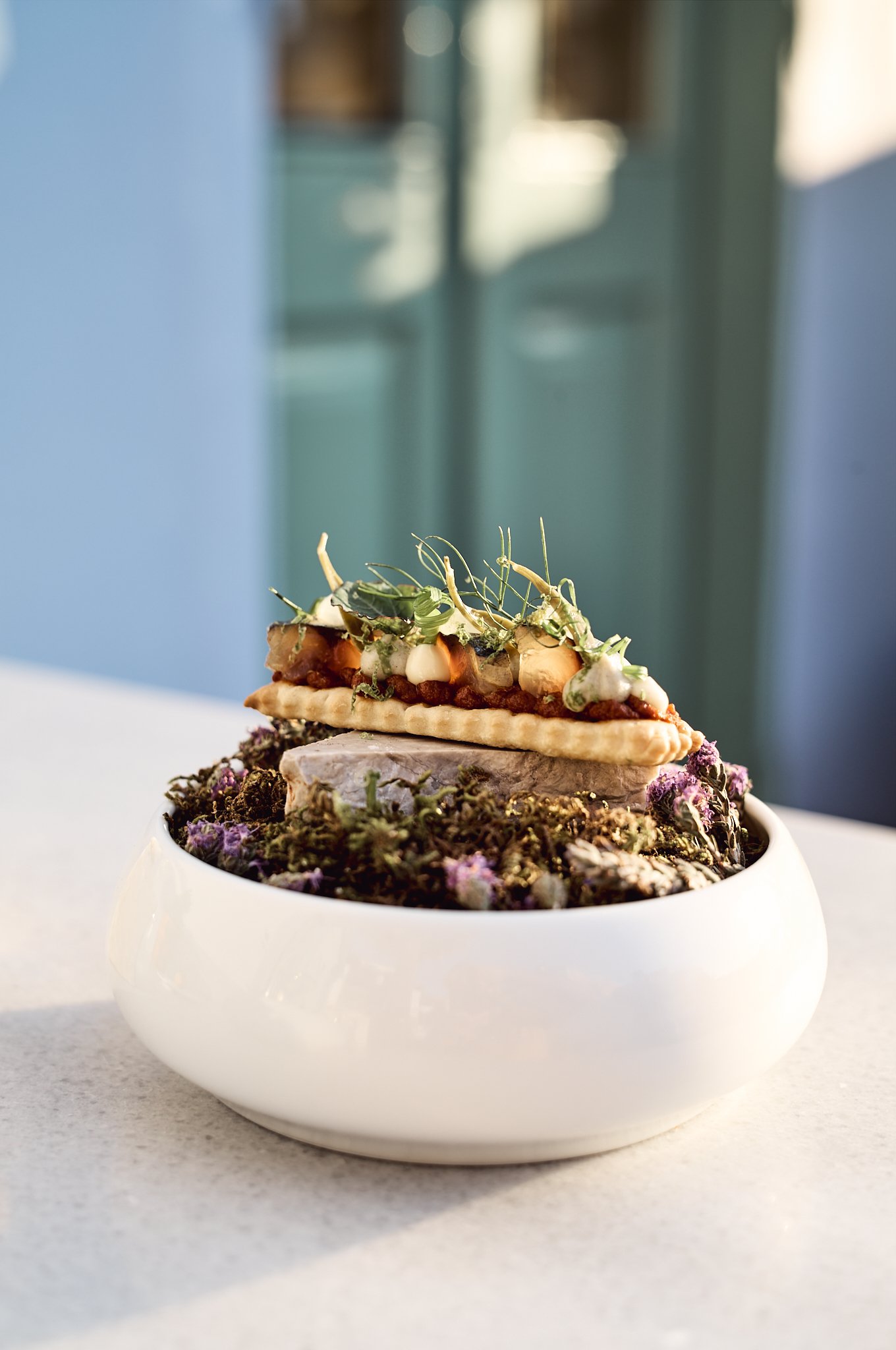 A decorative food arrangement in a white bowl on a white surface, featuring a layered arrangement of various ingredients including a pastry, vegetables or herbs, and a purple-tinted food item, with a softly blurred background. Best Restaurant in Symi