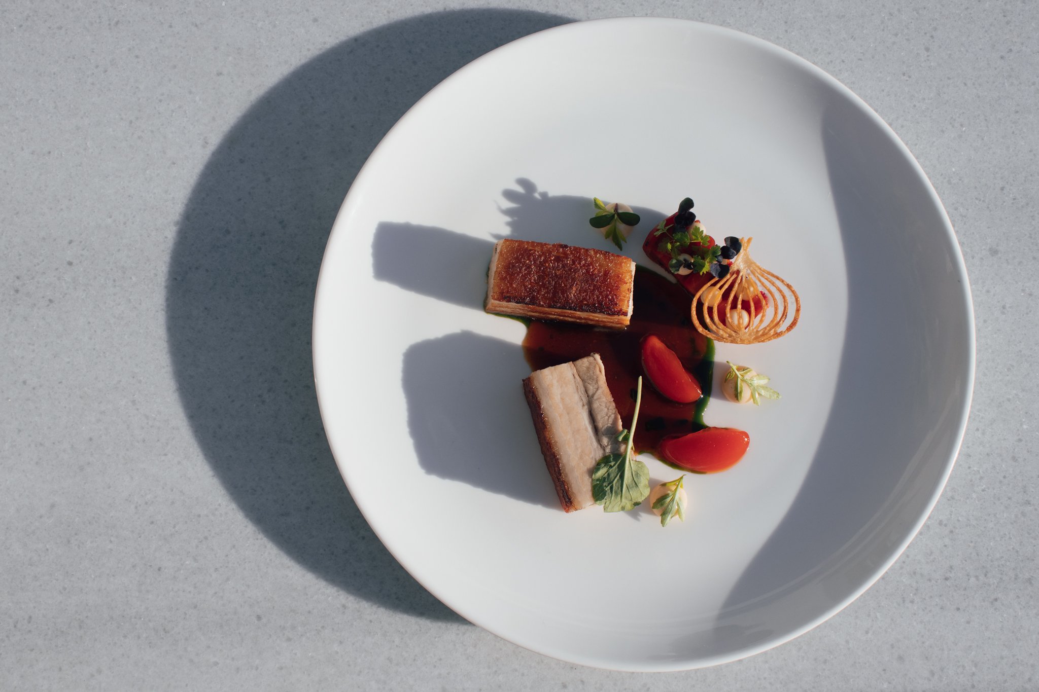Elegant plated dish with two pieces of meat, garnished with sauces, edible flowers, and microgreens, served on a white dish on a gray surface.