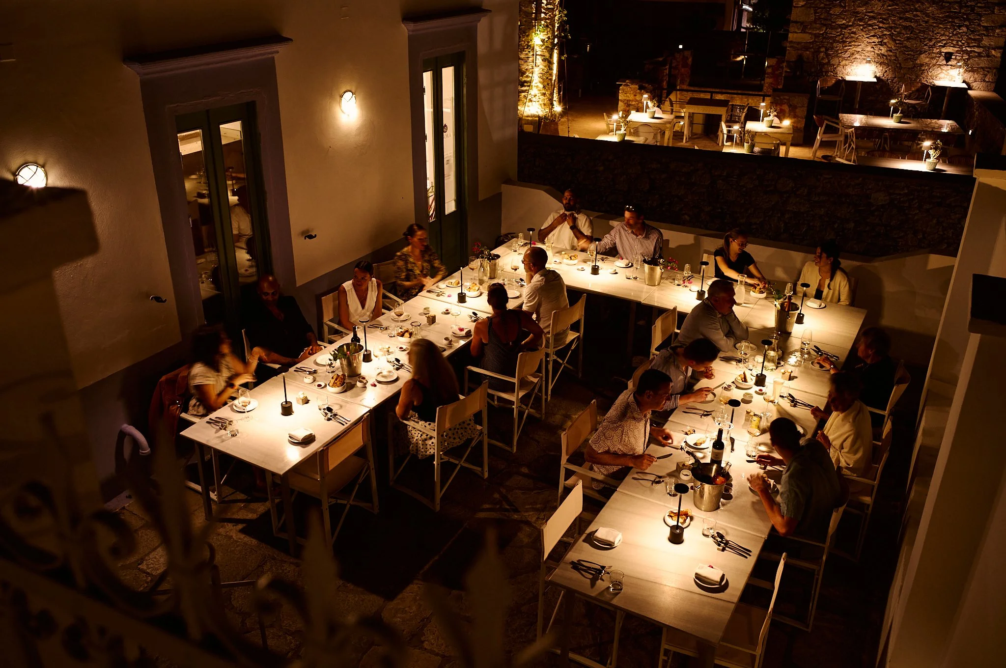 Events, weddings, birthdays, Baptisms, Agora Restaurant Symi. People dining at a restaurant during nighttime, sitting around illuminated tables with wine and food.