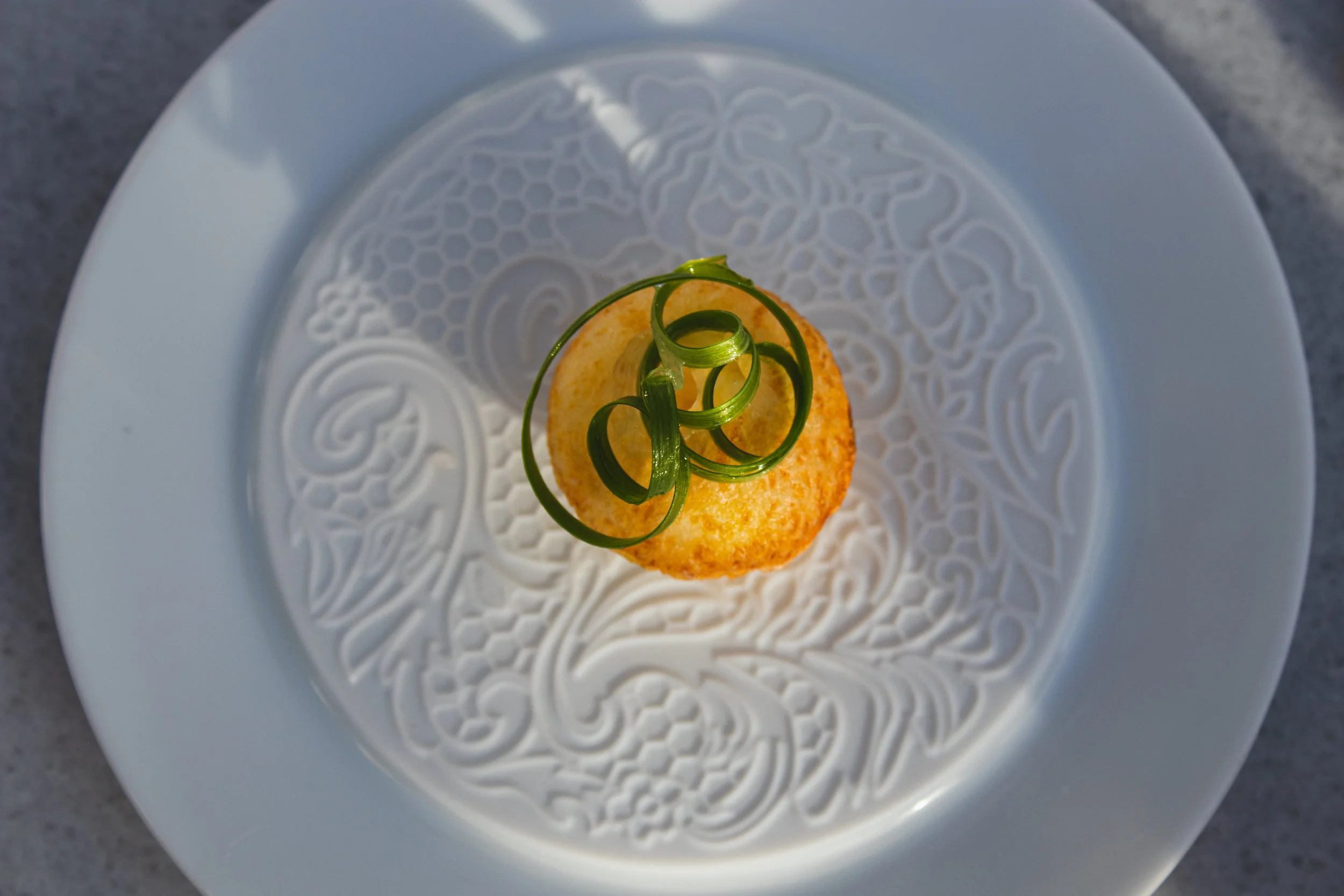 A round, golden-brown croquette garnished with curly green herb on a white plate with an intricate, embossed pattern. Gastronomy, Fine Dining, Symi, Greece