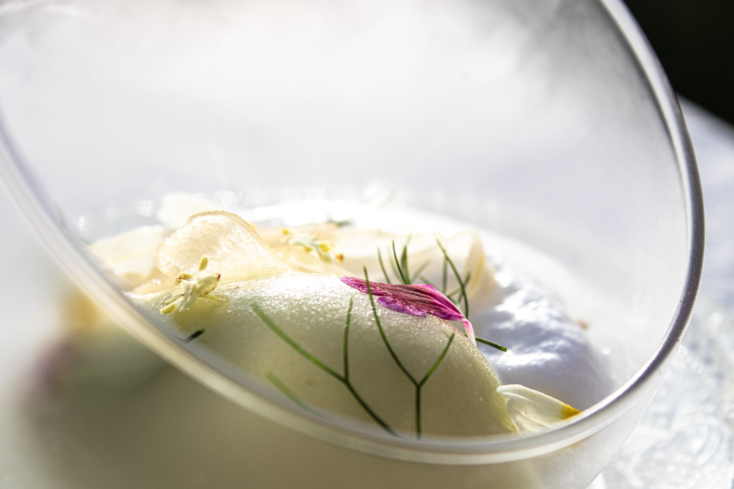 Close-up of a wedding cake with white icing, garnished with flower petals and small edible flowers, inside a clear plastic cake container.