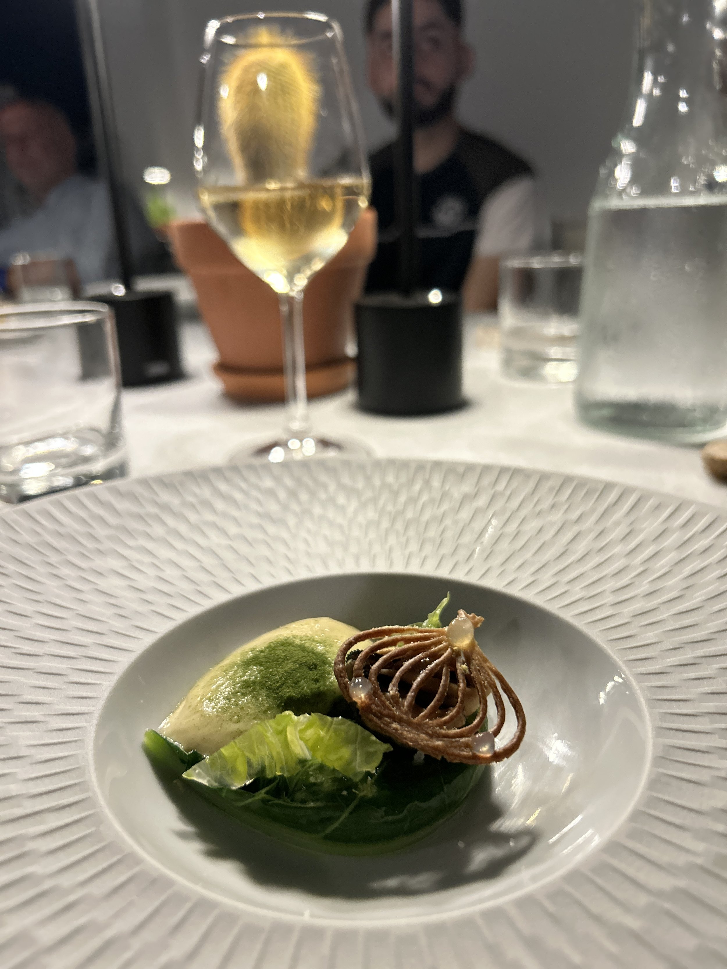 A gourmet dish with a green vegetable, a dollop of green sauce, and a decorative spiral-shaped element on a white textured plate. In the background, there is a glass of white wine, a water glass, and other drinkware on a white tablecloth, with blurre