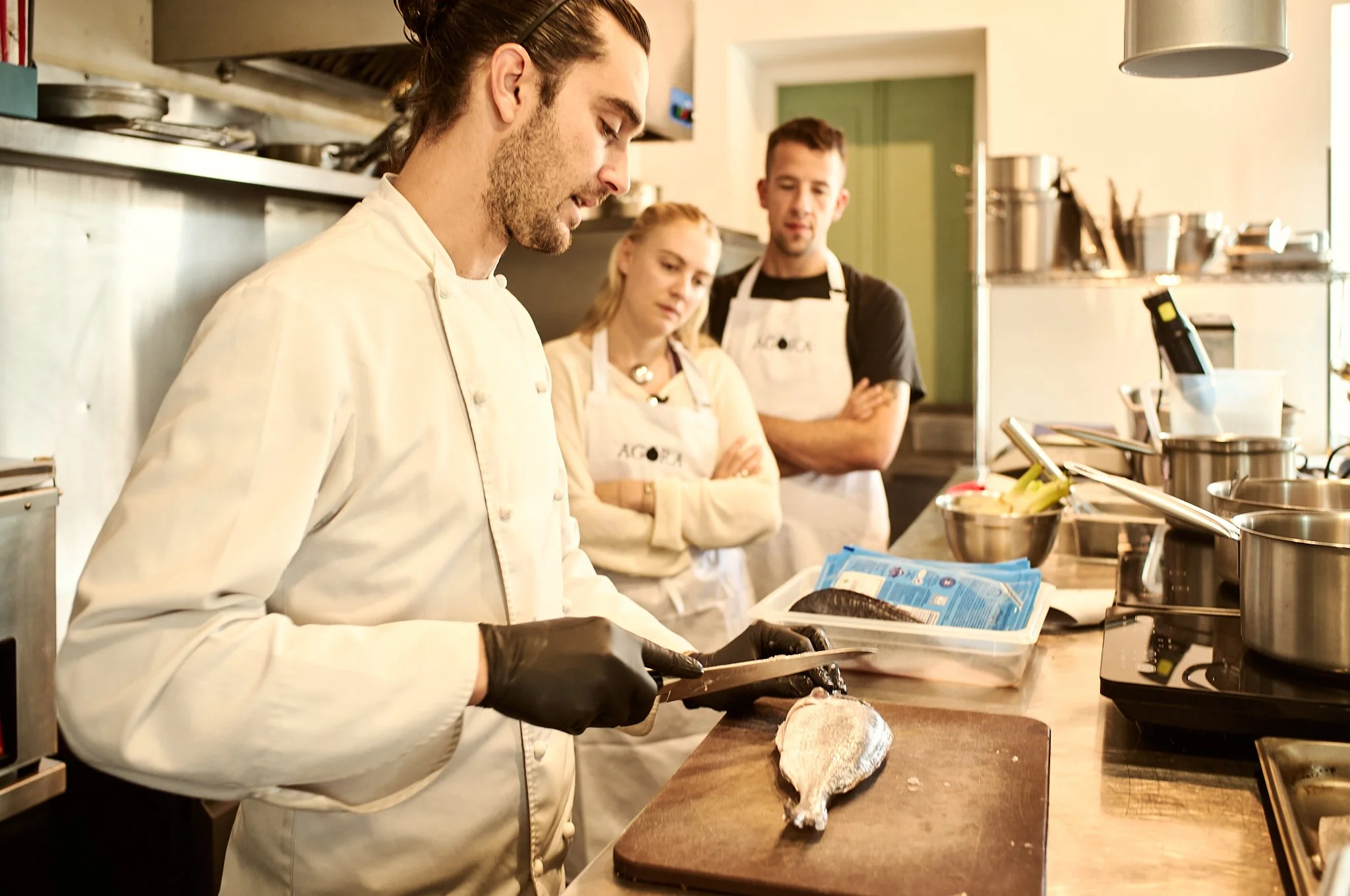 Masterchef Greece Chef Chris, is slicing fish on a cutting board in a professional kitchen, with two people observing in the background.