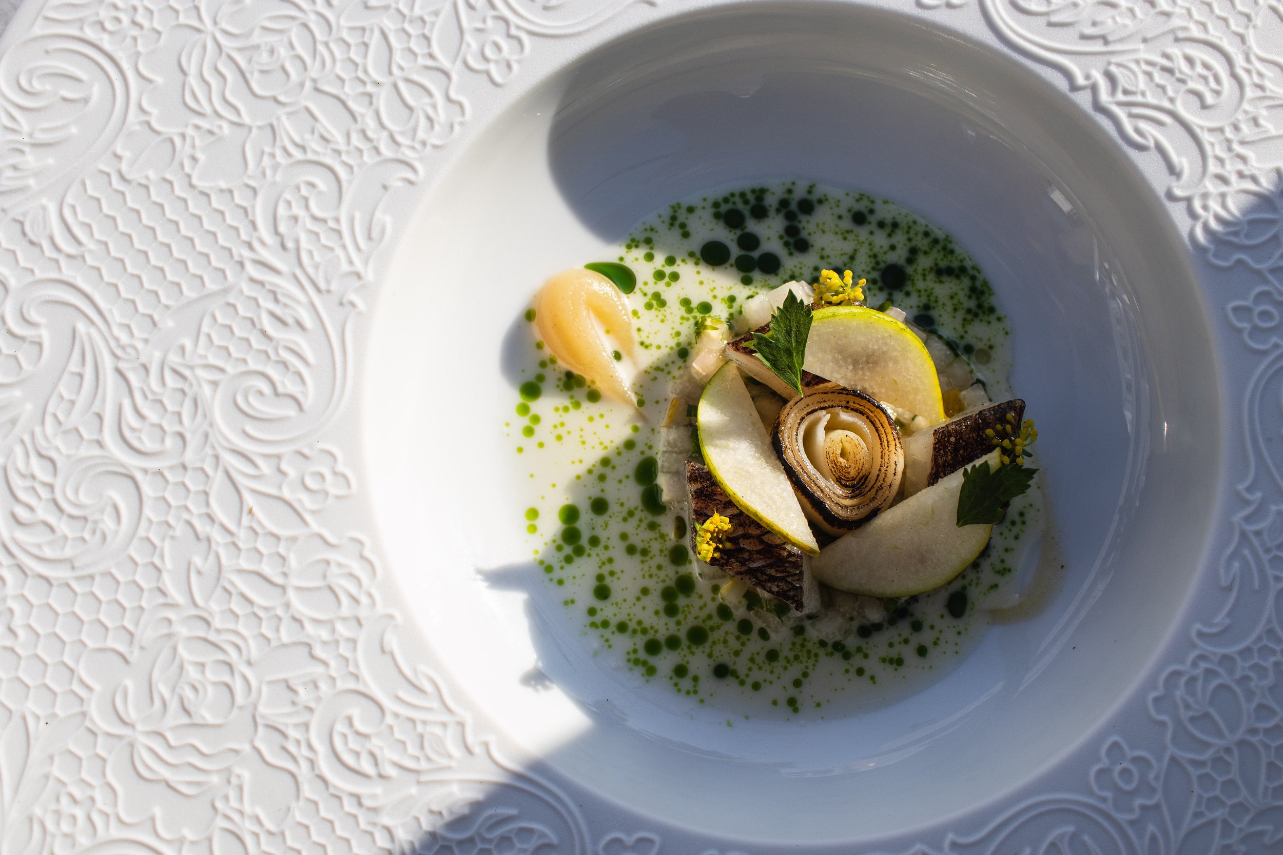 A white bowl with intricate embossed pattern on the rim, containing a gourmet dish with sliced pears, a rolled slice of cured meat or fish, green sauce, and garnished with edible flowers and herbs.