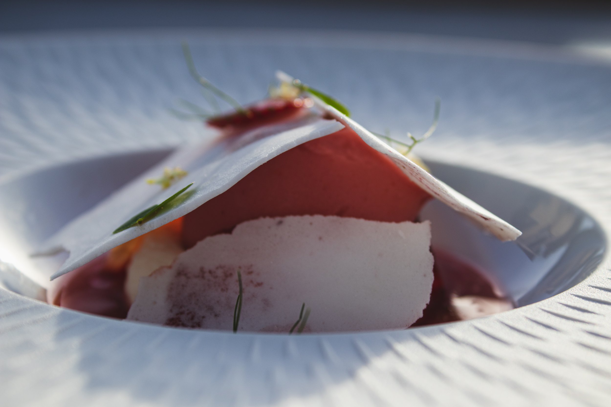 Close-up of a plated dessert with pink mousse, white chocolate shards, and garnishes, inside a textured white plate.