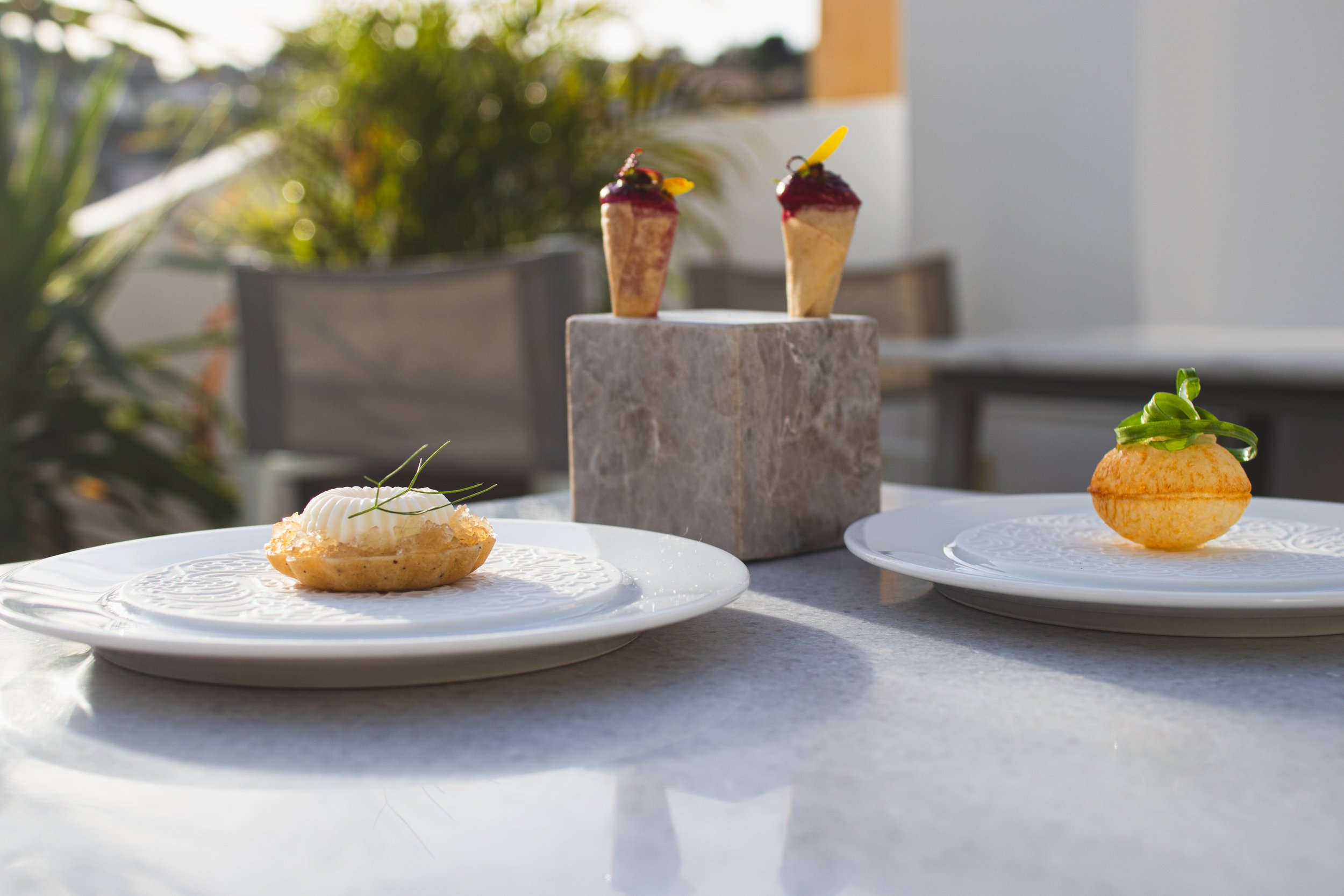 Elegant dessert plates with tart and mousse, and two cone-shaped pastries with berry topping on a marble stand, set on a white table outdoors.