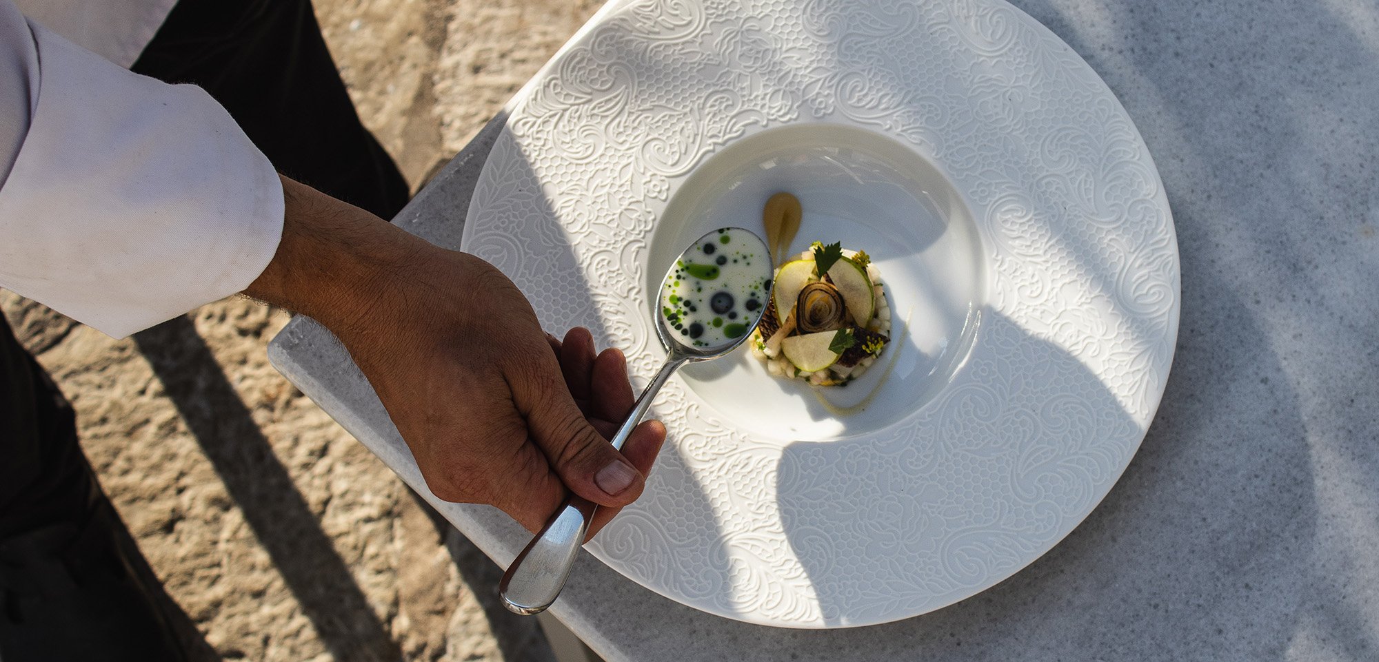 A chef's hand holding a spoon with a creative dessert, featuring mousse, chocolate, and garnishes, served on an ornate white plate with intricate detailing, placed on a white surface outdoors.