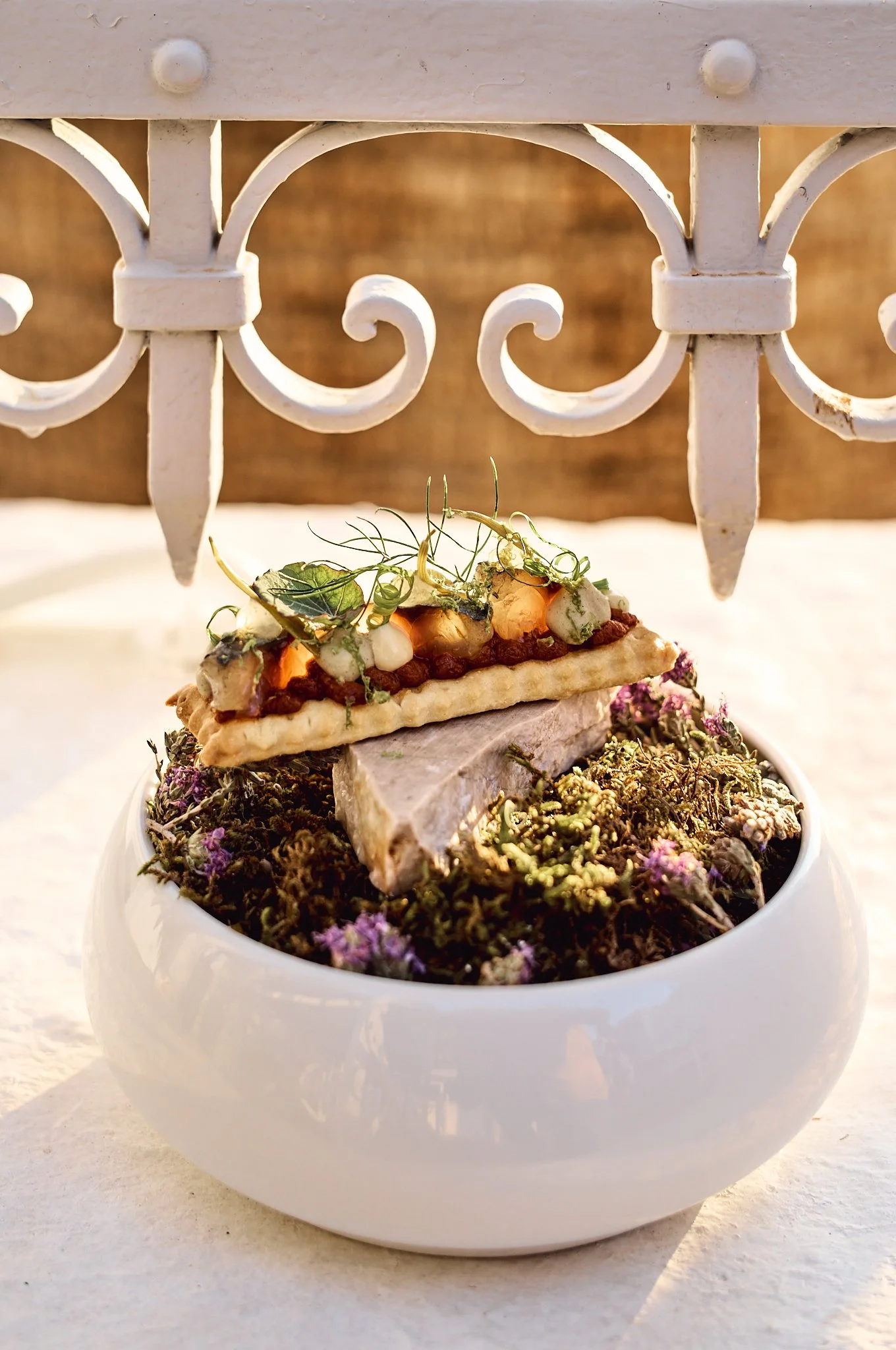 Decorative dish with fish, garnished with herbs and vegetables, placed in a white bowl on a table, with a white decorative fence in the background. From Symi's finest restaurant, Agora.