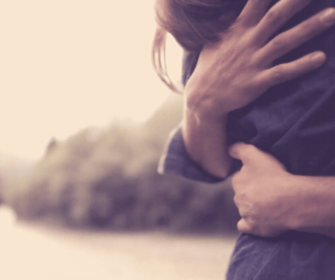 A person man hugging a woman, set outdoors in front of a lake and mountains, with a blurred background.