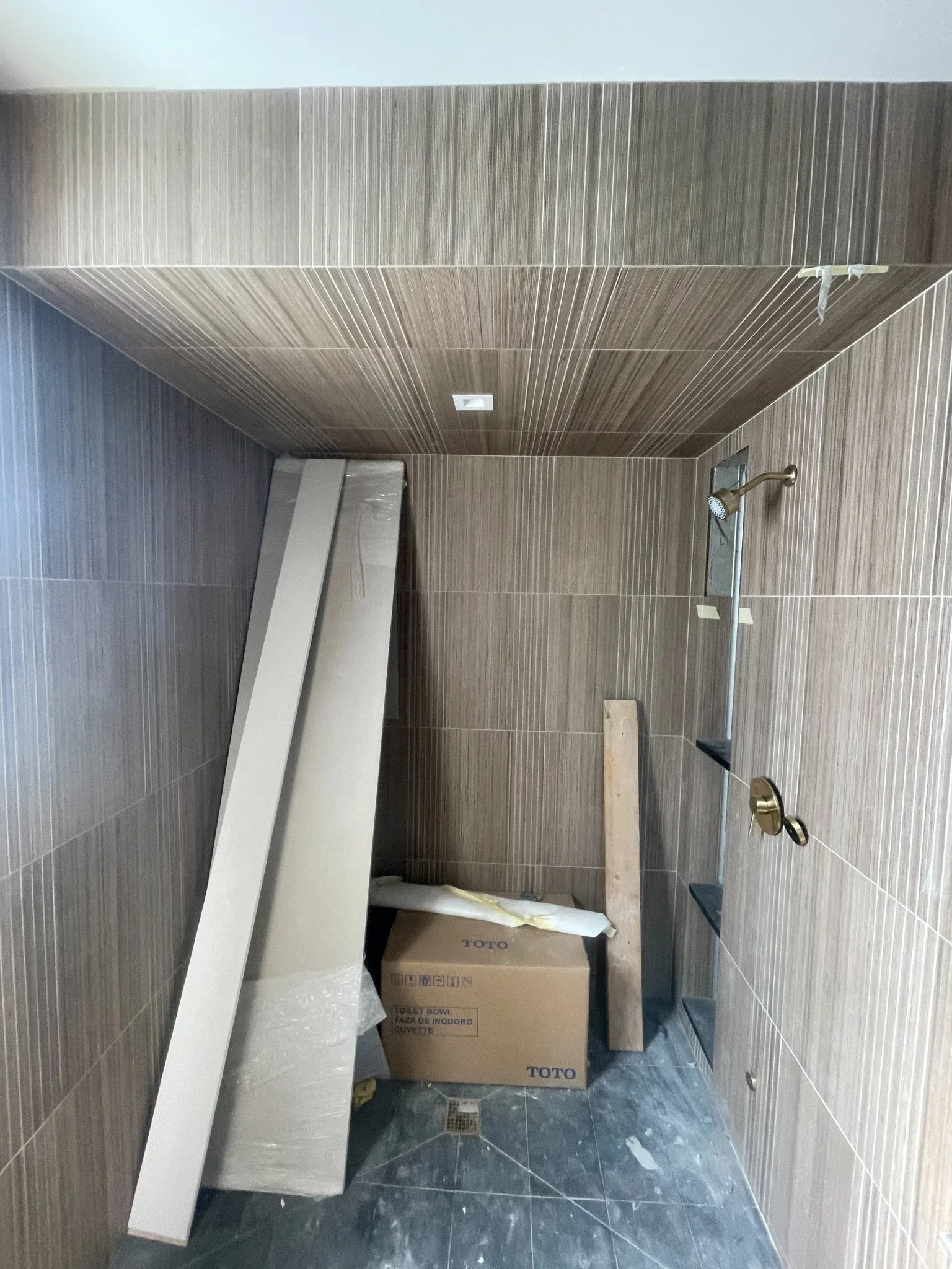 This image shows a small bathroom under construction with brown tiled walls and a dark tile floor. There is a cardboard box labeled 'TOTO' and some construction materials leaning against the wall, including a piece of wood and a cardboard panel. An u