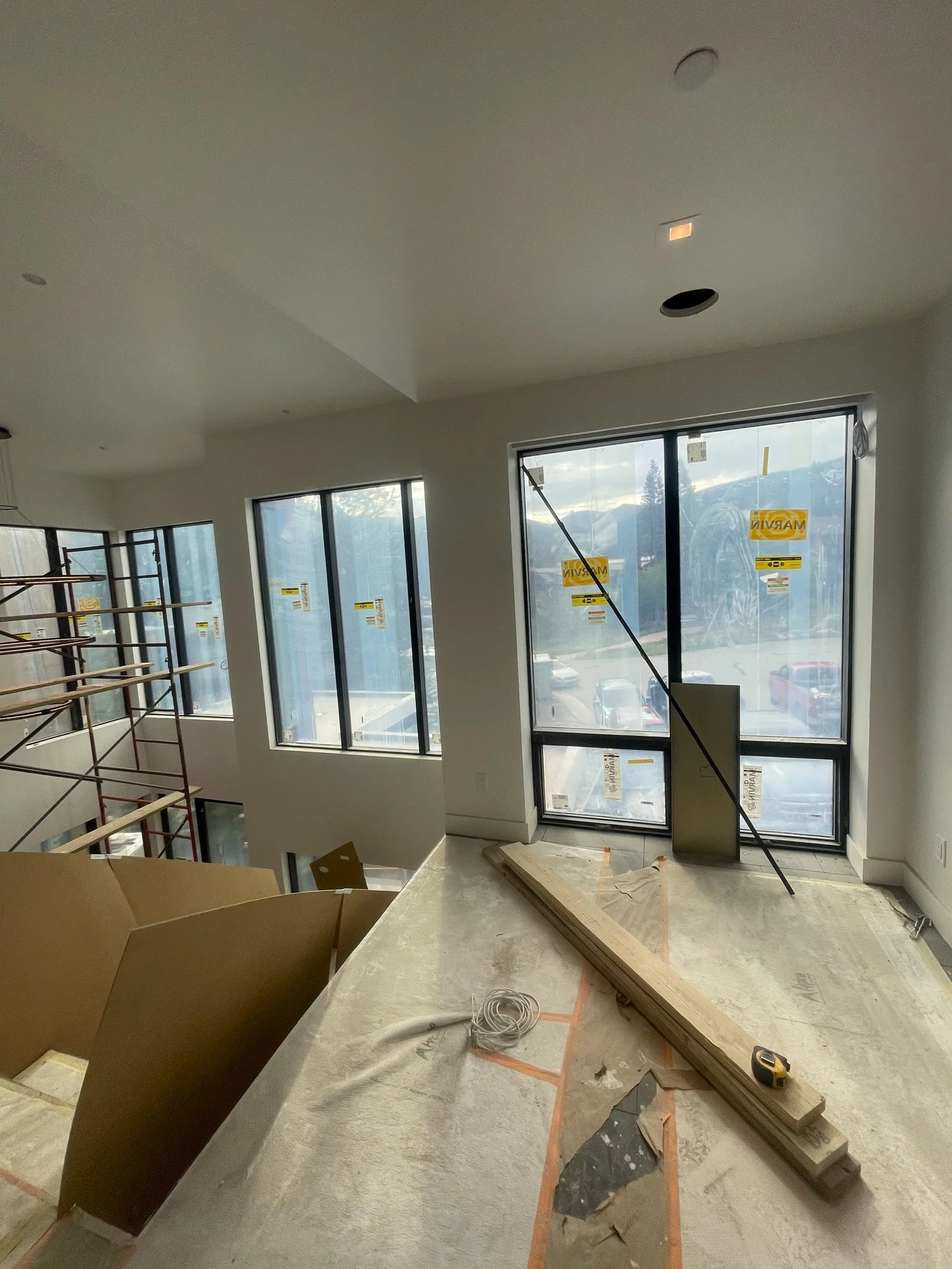 Interior view of a building under construction with large windows, construction materials, and tools on the floor.