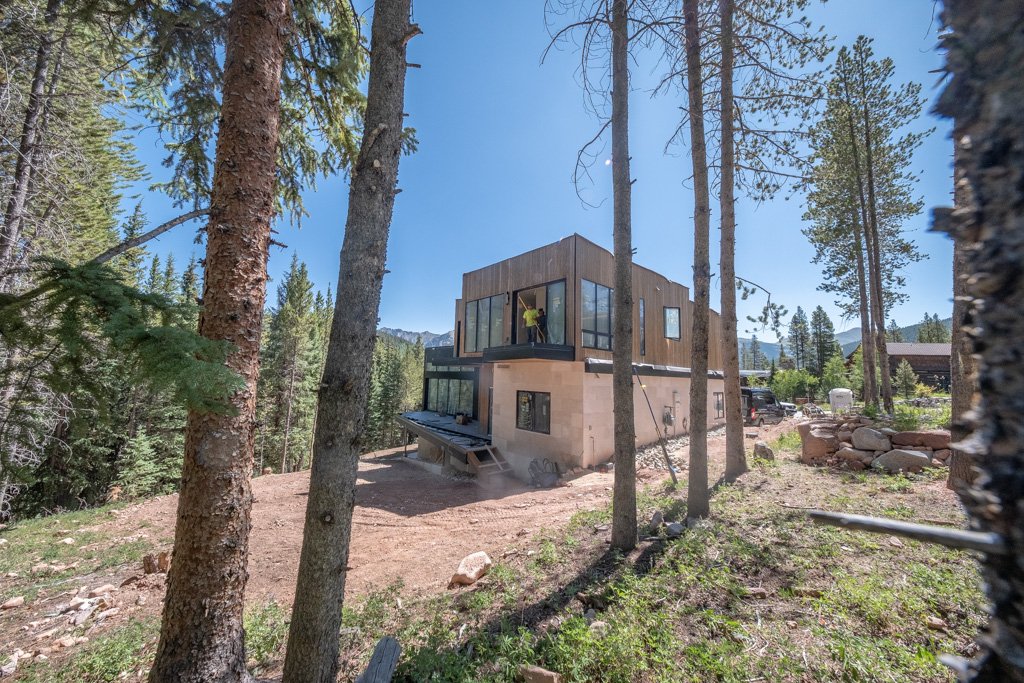 Construction of a modern two-story house in a forested area with tall pine trees, mountains in the background, and construction workers working on the upper level.
