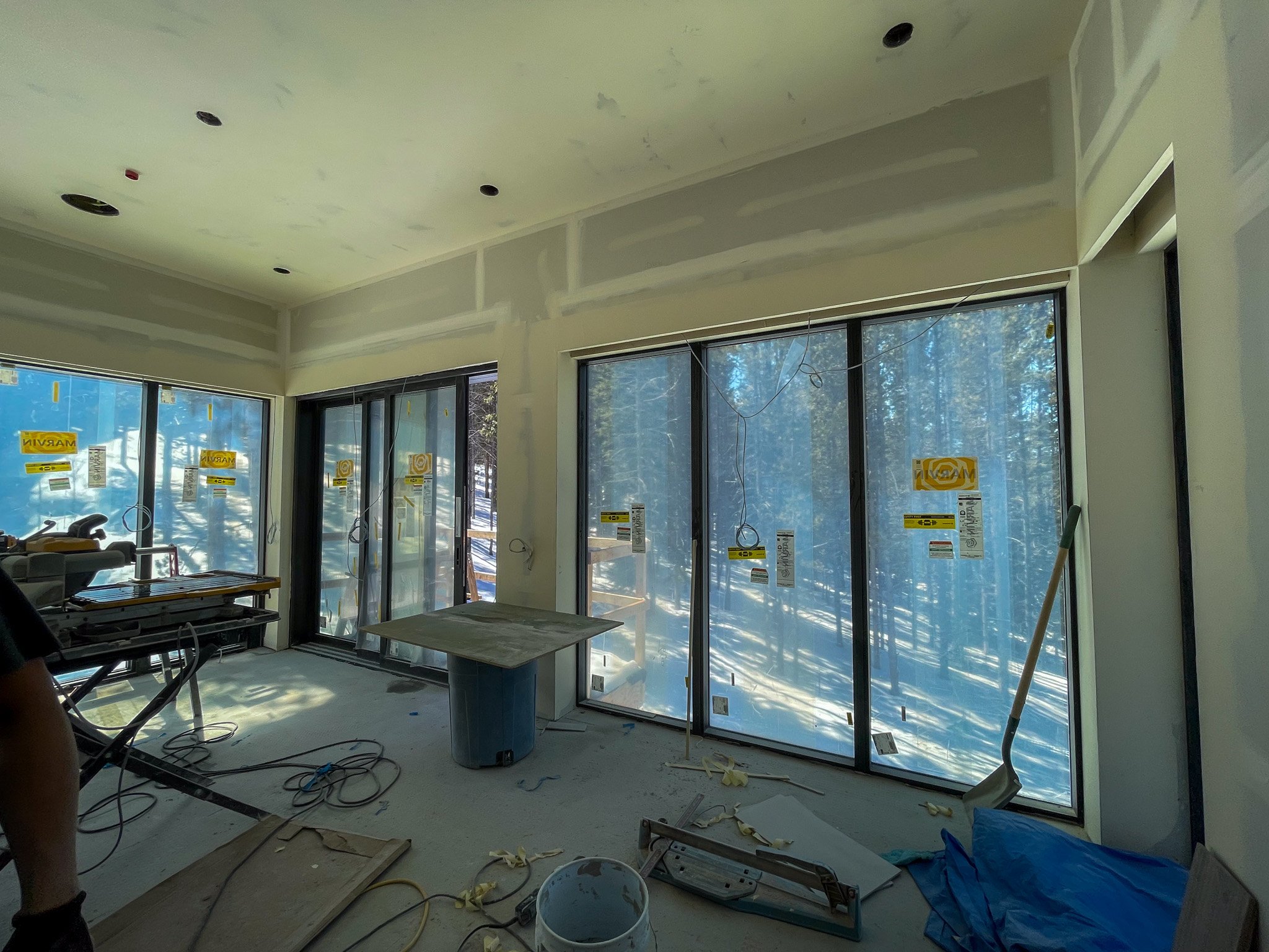 Interior view of a room under construction with large windows, visible wiring, construction tools, and materials scattered on the floor.