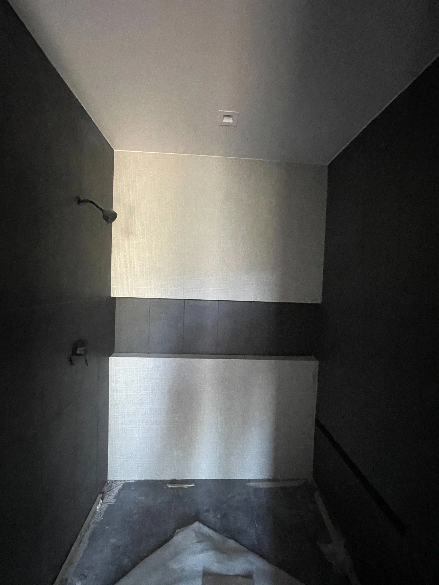 Unfinished shower with dark walls, a showerhead, and a lower panel with a textured light-colored surface, with some construction materials on the floor.
