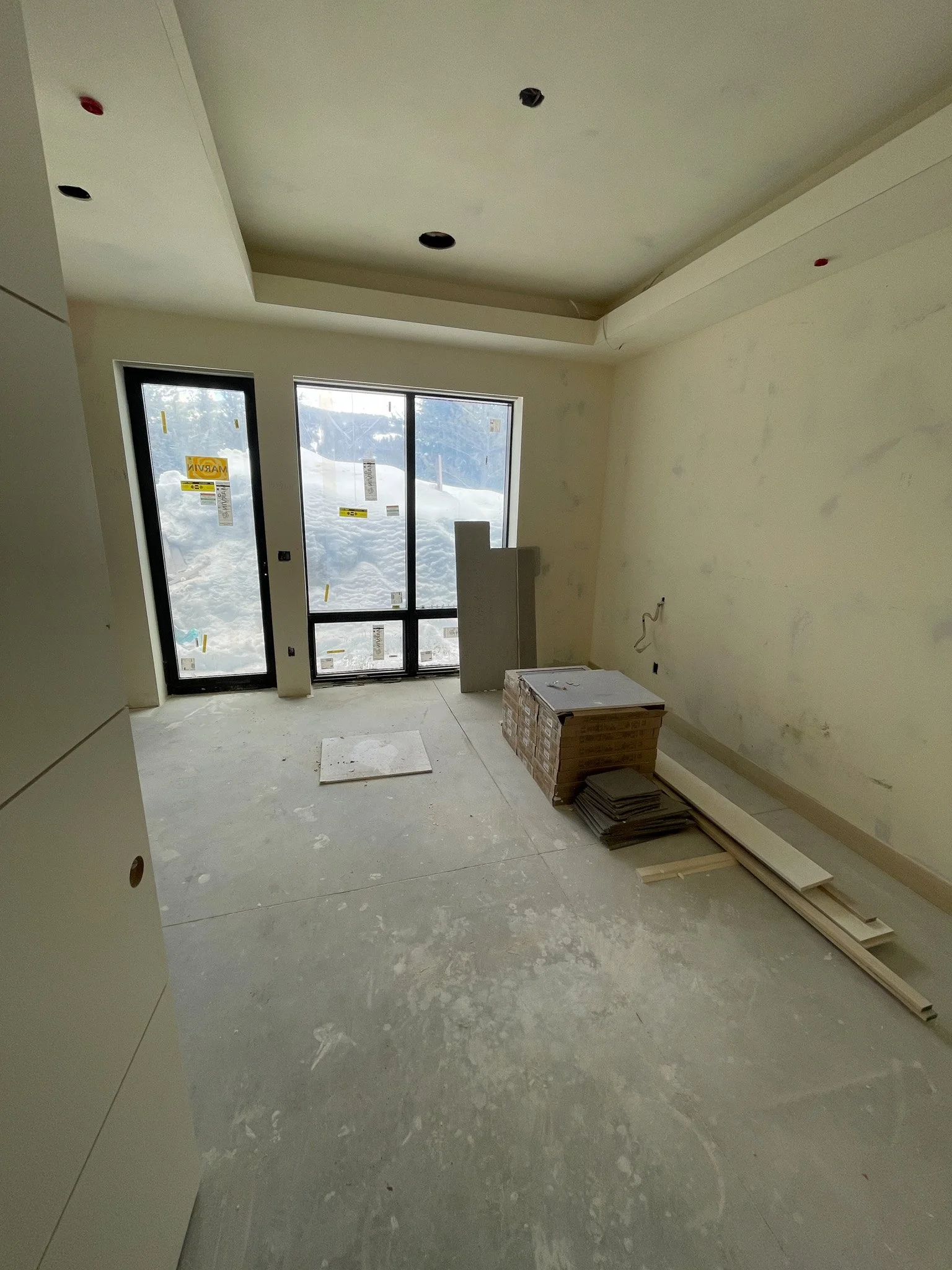 An interior room under construction with large windows showing snowy outdoor scenery, unfinished walls, and construction materials scattered on the floor.