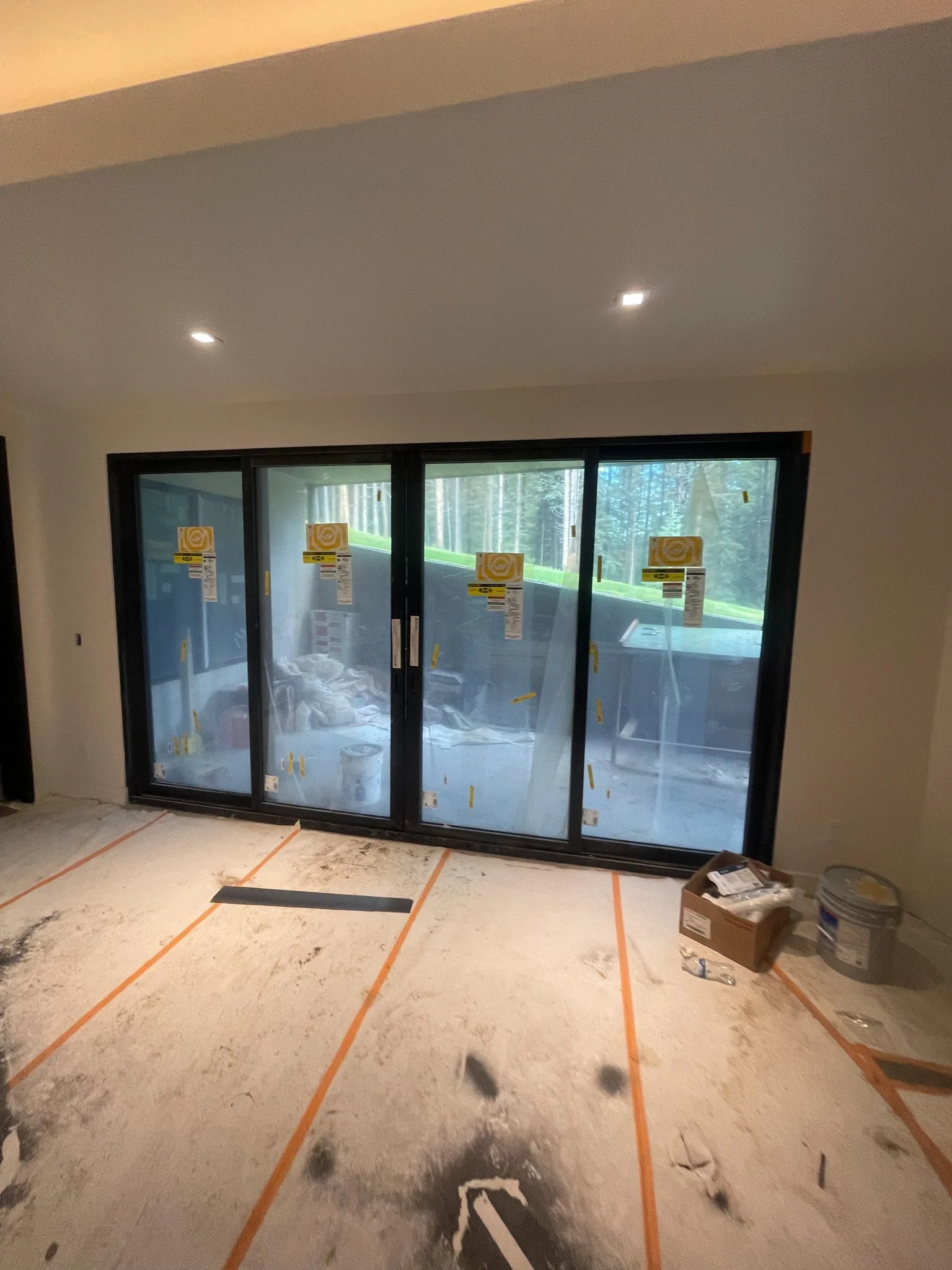 Inside a room under construction, with sliding glass doors covered in protective film and paper, construction tools and supplies on the floor, and a view of a forest outside.
