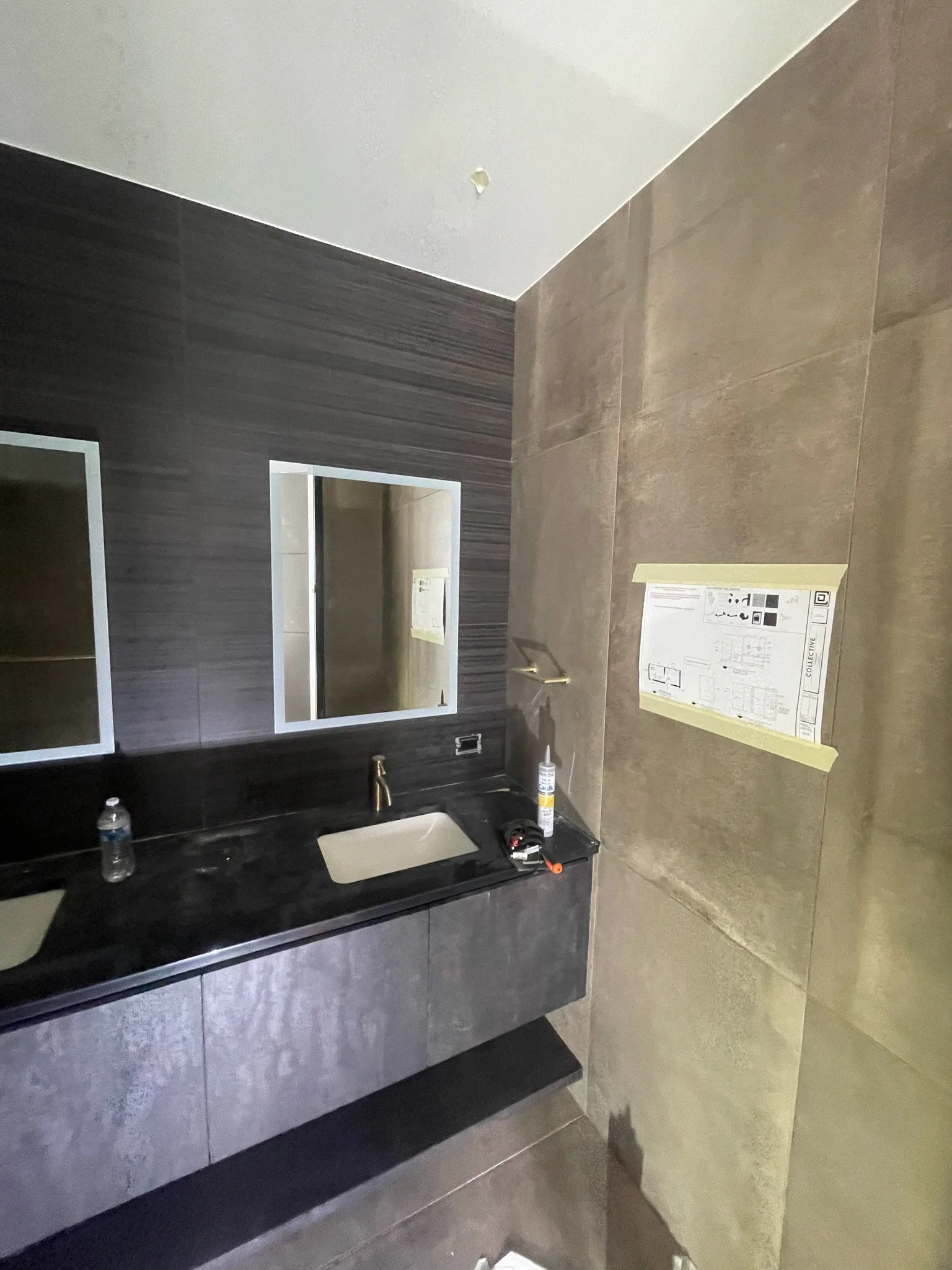 A bathroom under construction with a black countertop, a white sink, a mirror, and gray tiled walls.