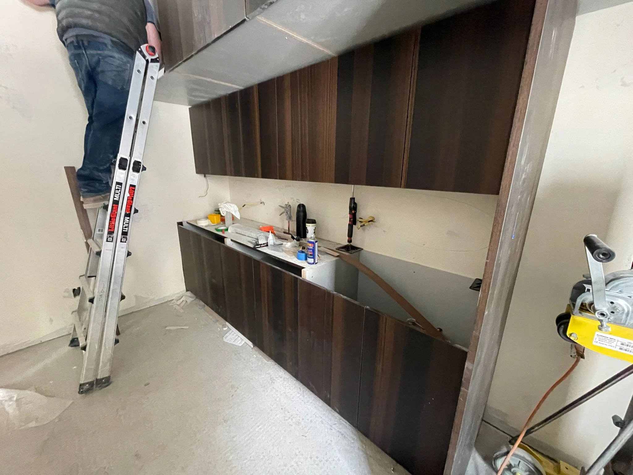 Kitchen under renovation with dark wooden cabinets, construction tools, and a worker on a ladder installing cabinetry.