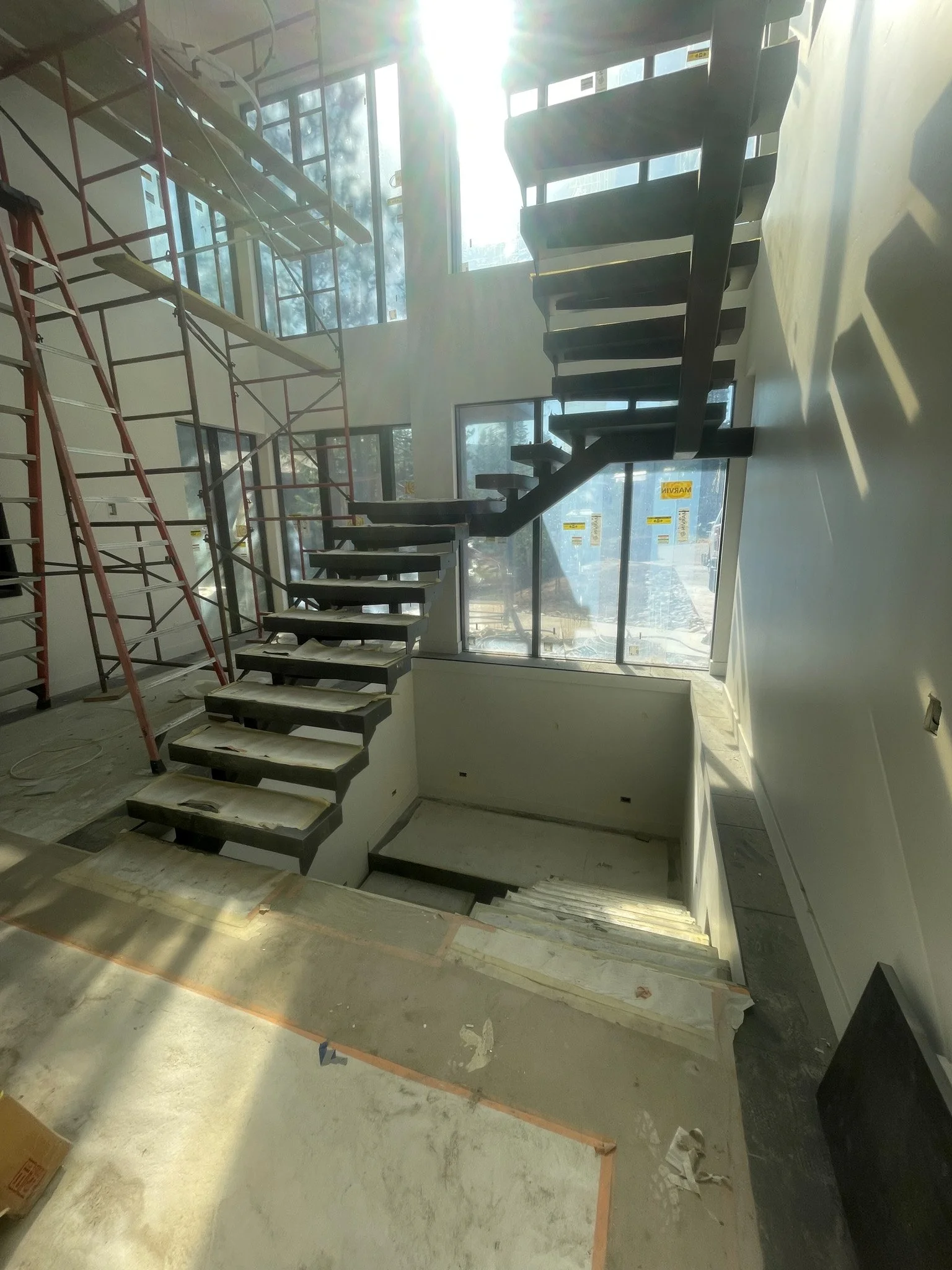Interior of a building under construction with a staircase and scaffolding, sunlight streaming through large windows.