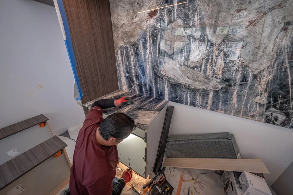 A person working on installing a dark, patterned marble countertop in a kitchen under construction.