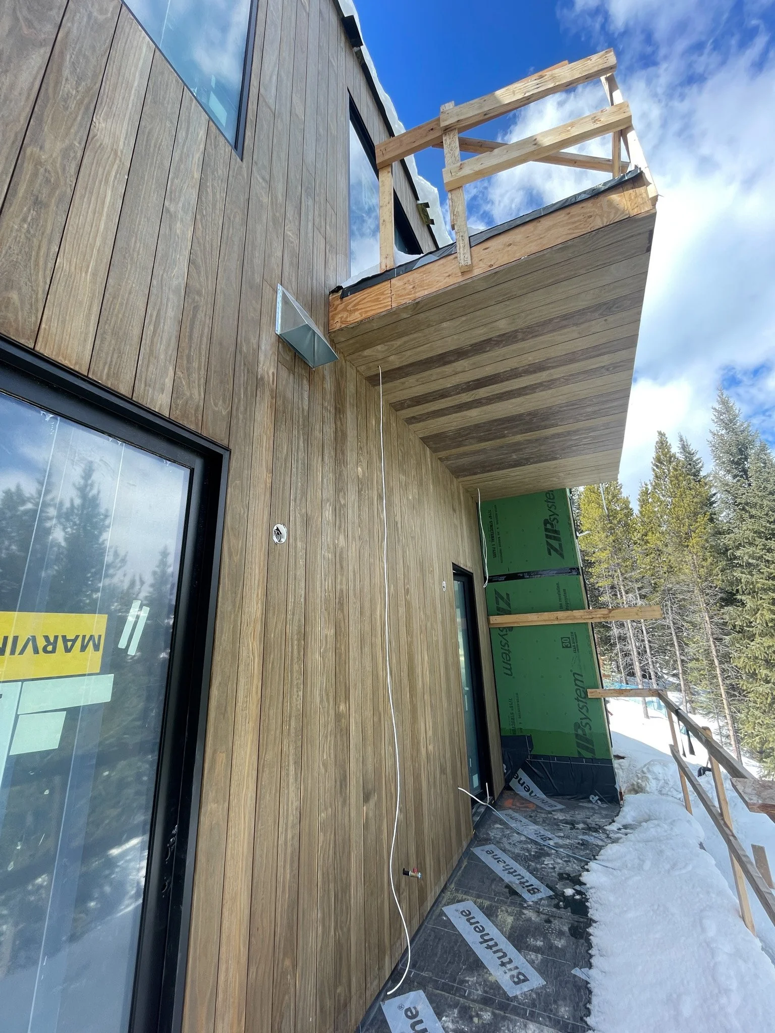 New wooden deck on a house under construction, with a balcony and snow on the ground, surrounded by trees and blue sky.