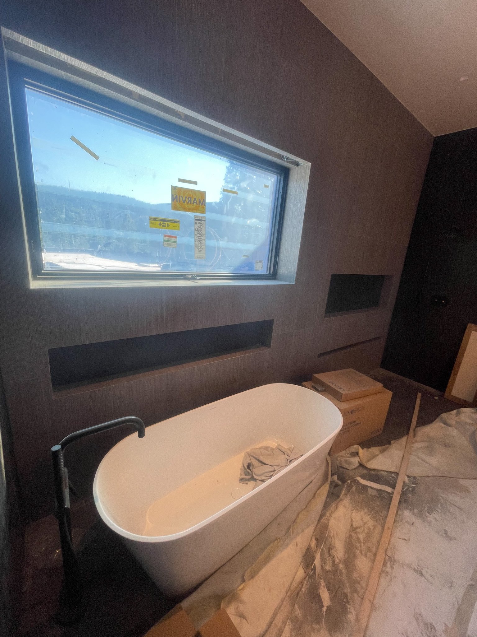 A bathroom under construction with a white freestanding bathtub, a black faucet, a large window, and a partly finished wall with built-in shelves. Construction materials and tools are scattered on the floor.