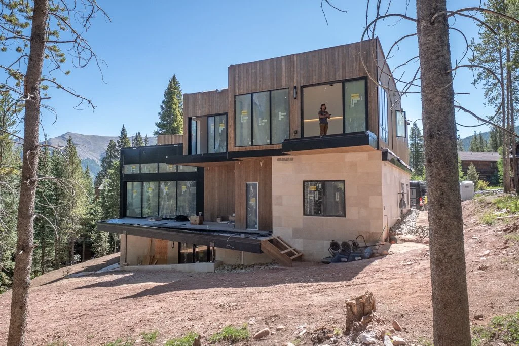 A modern two-story house under construction in a forested area, with large glass windows and wood exterior panels.