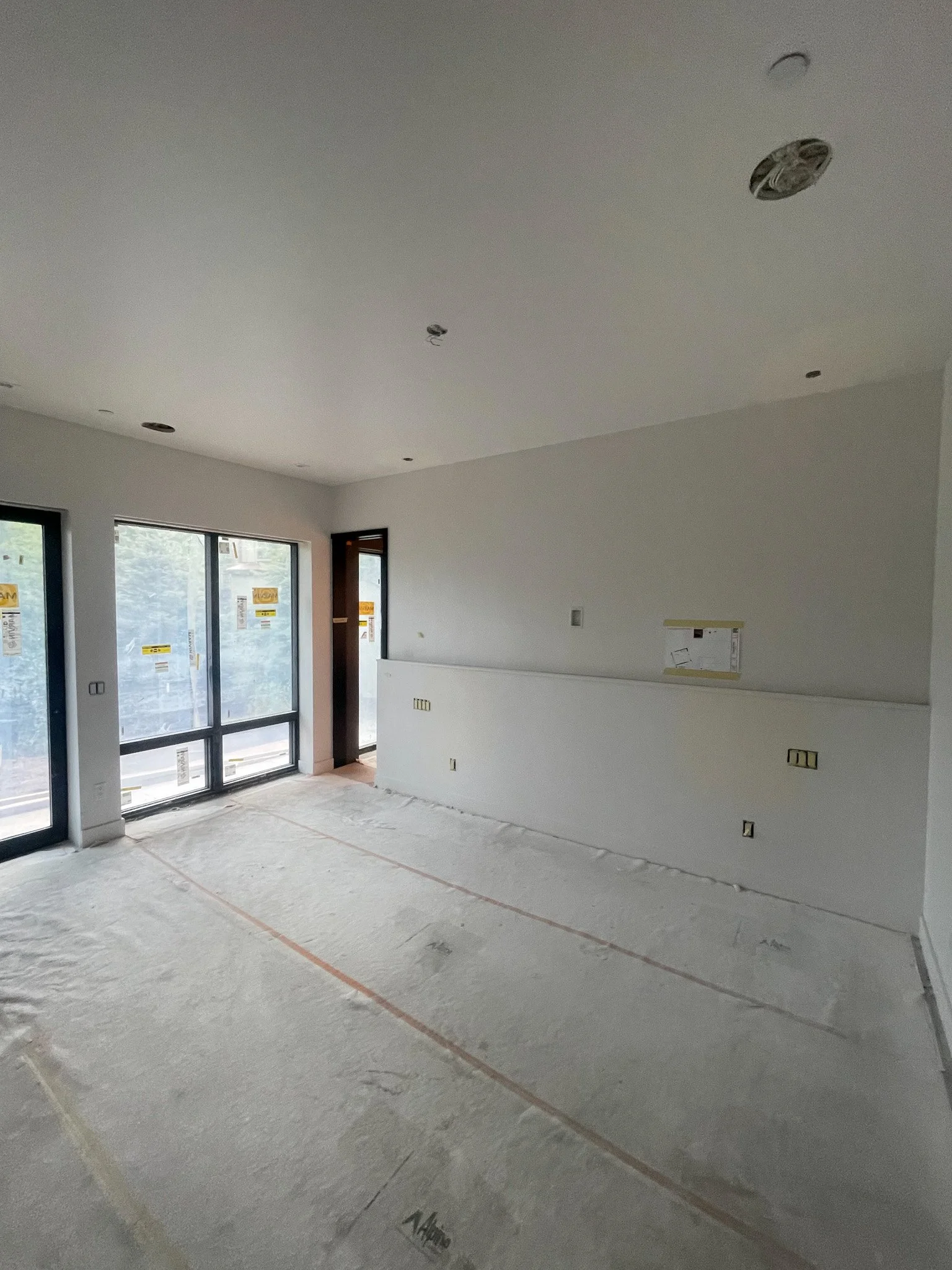 An unfinished interior room under construction with white walls, large glass sliding doors, and a partially installed ceiling with recessed lighting fixtures.
