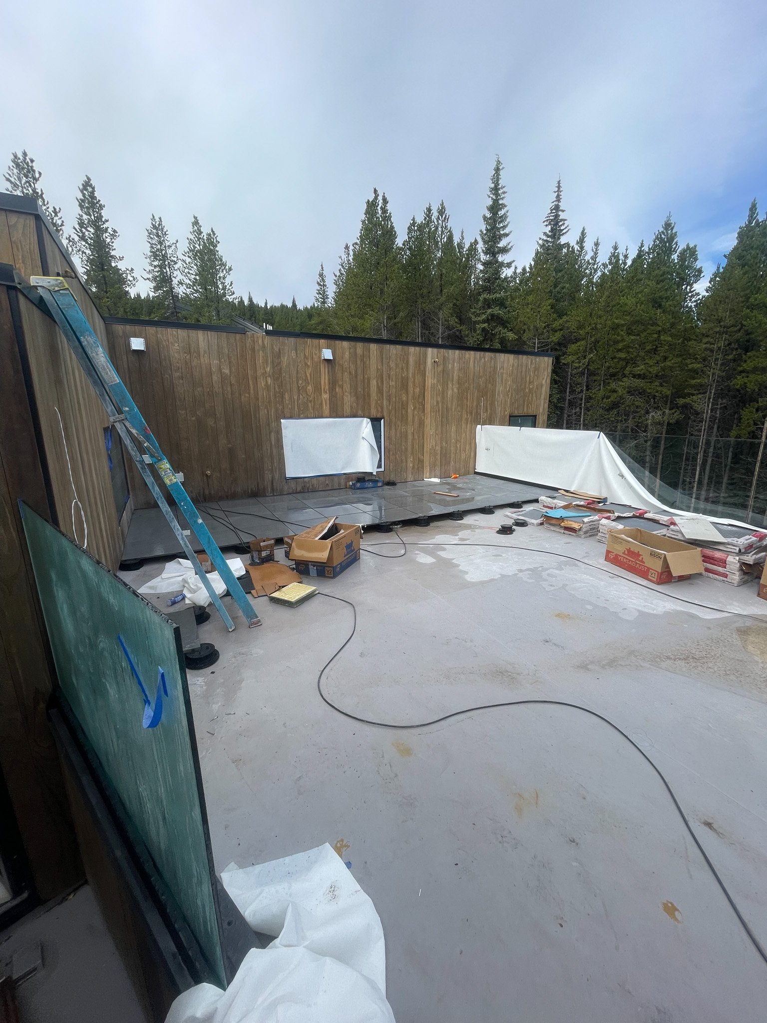 Construction site on a terrace with tools, boxes, and partially installed glass or windows, surrounded by a wooden fence and trees in the background, under a cloudy sky.