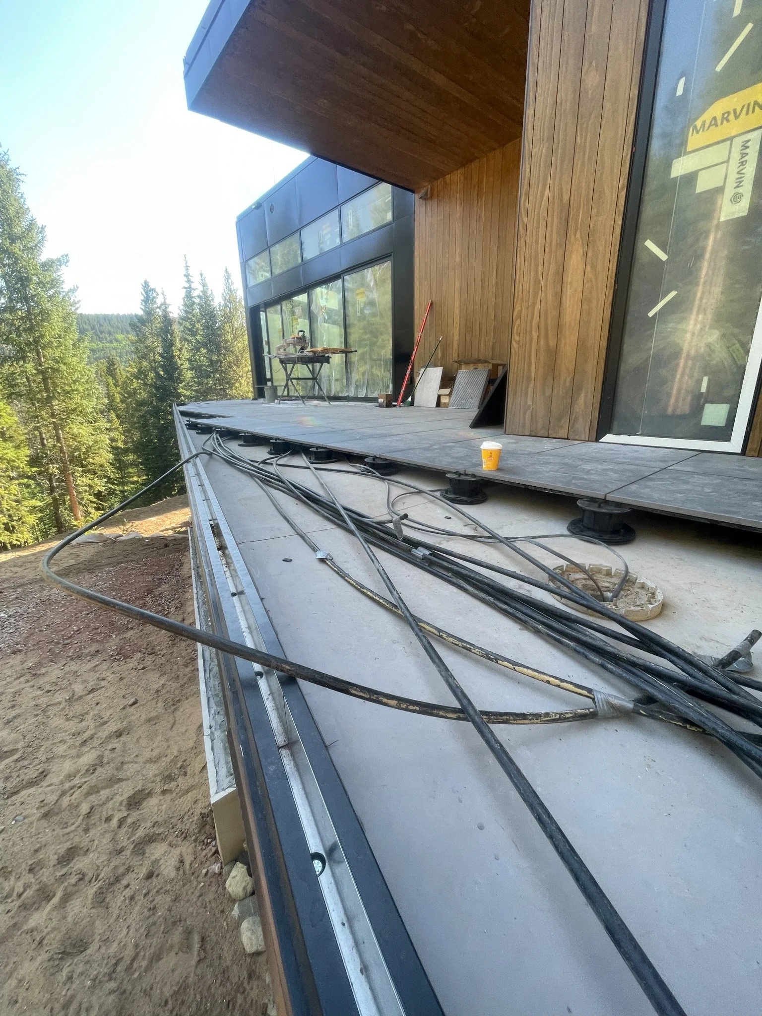 Construction site on a hillside with a modern building, wires and cables on the ground, and construction tools and materials visible on the deck.