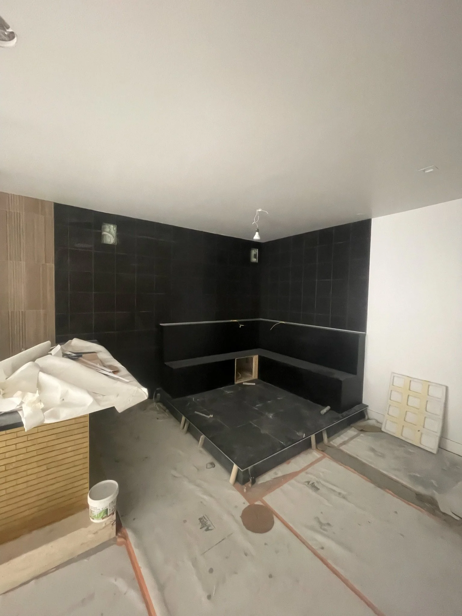Interior of a room under construction with a black tiled wall, partially installed flooring, and construction materials scattered on the floor.