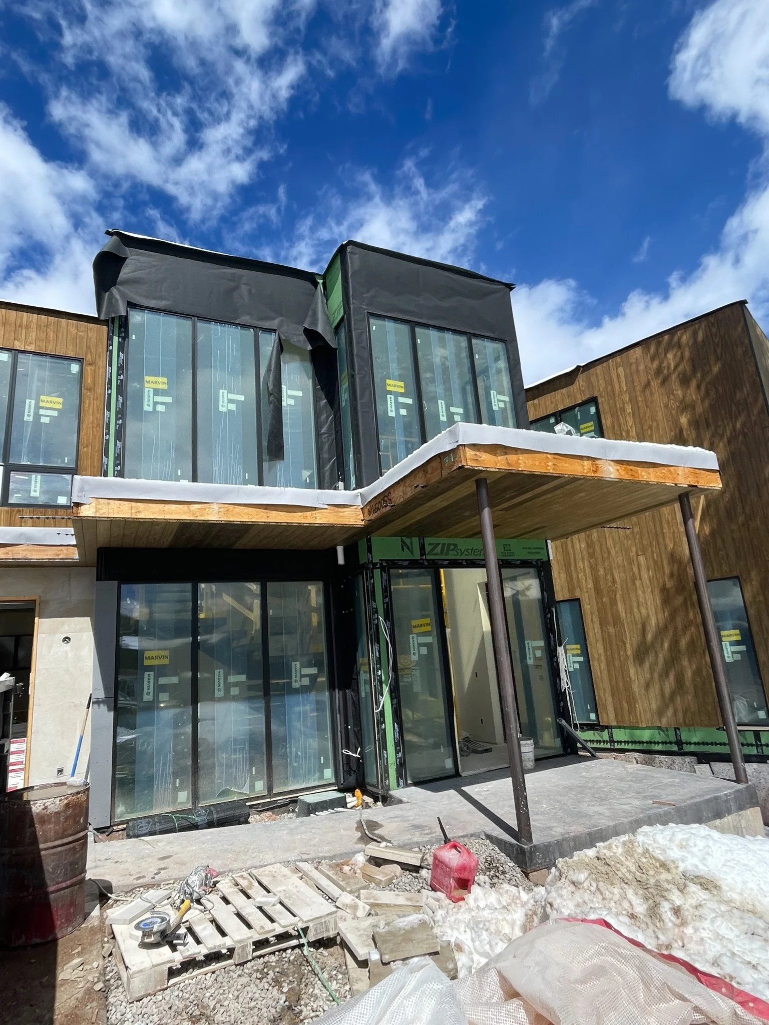 Construction site of a modern building with large glass windows and wood paneling, partially finished with some snow on the ledges, under a blue sky with clouds.