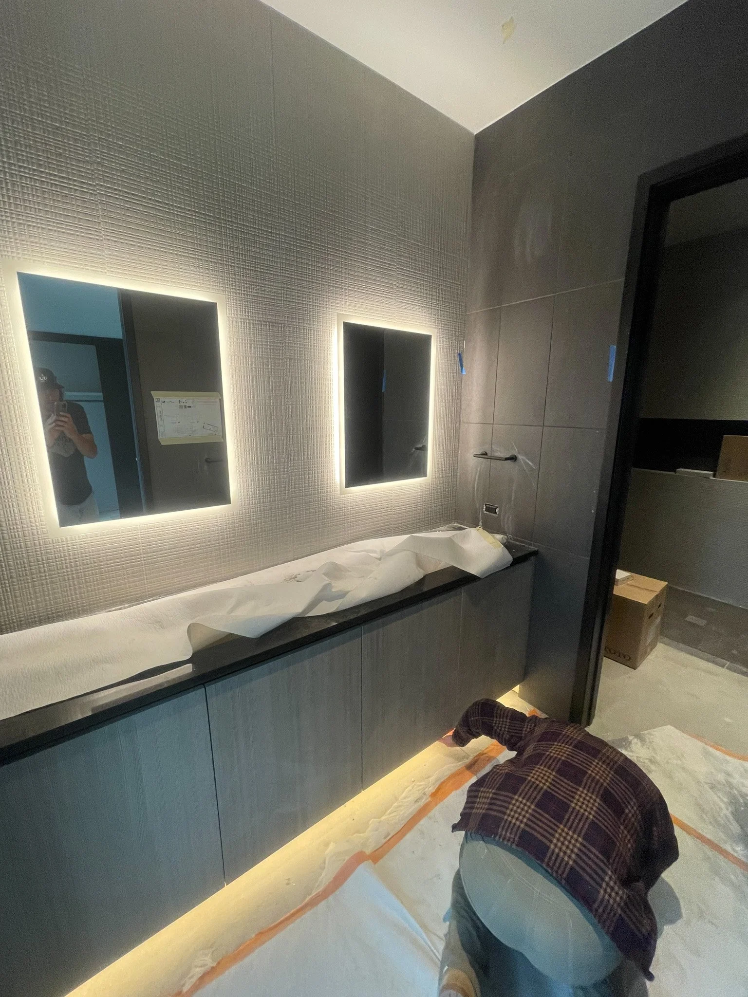 A person kneeling on the floor of a bathroom under construction, working on the cabinetry. The bathroom has textured wall panels, two illuminated mirrors above a black countertop, and lighting underneath the cabinets.