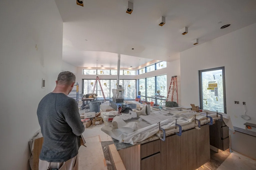 A man standing inside a house under construction, with tools, materials, and ladders, and new windows revealing a view of trees outside.