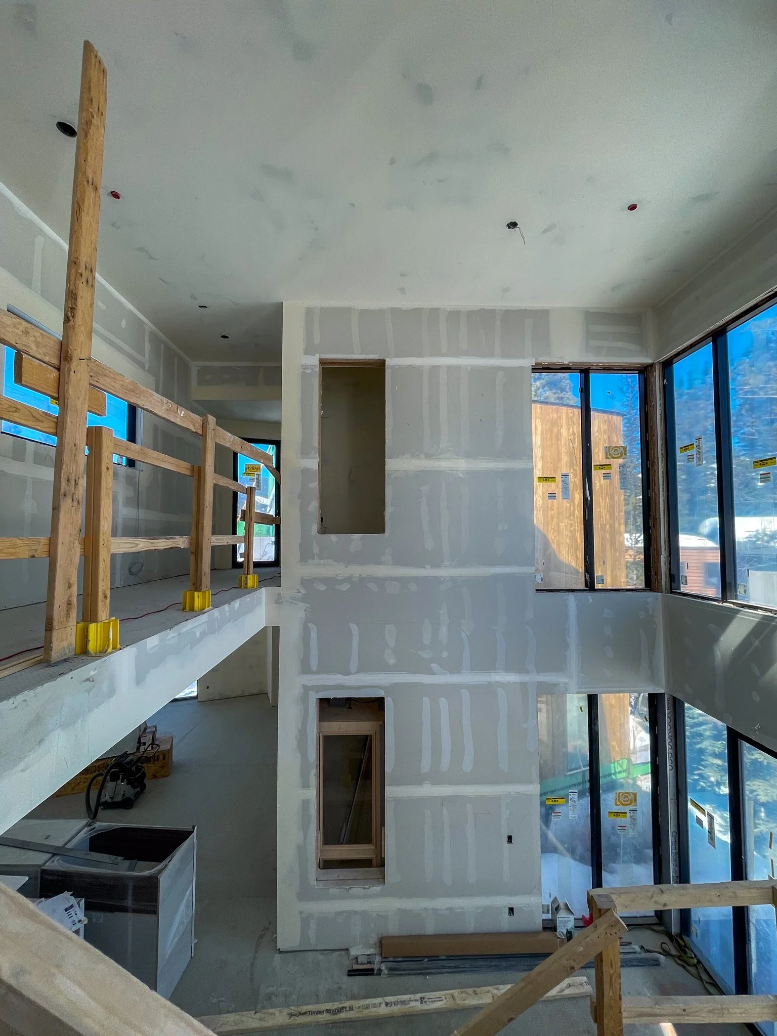 Interior view of a building under construction with drywall and large windows, showing an open space with unfinished walls and a wooden railing.