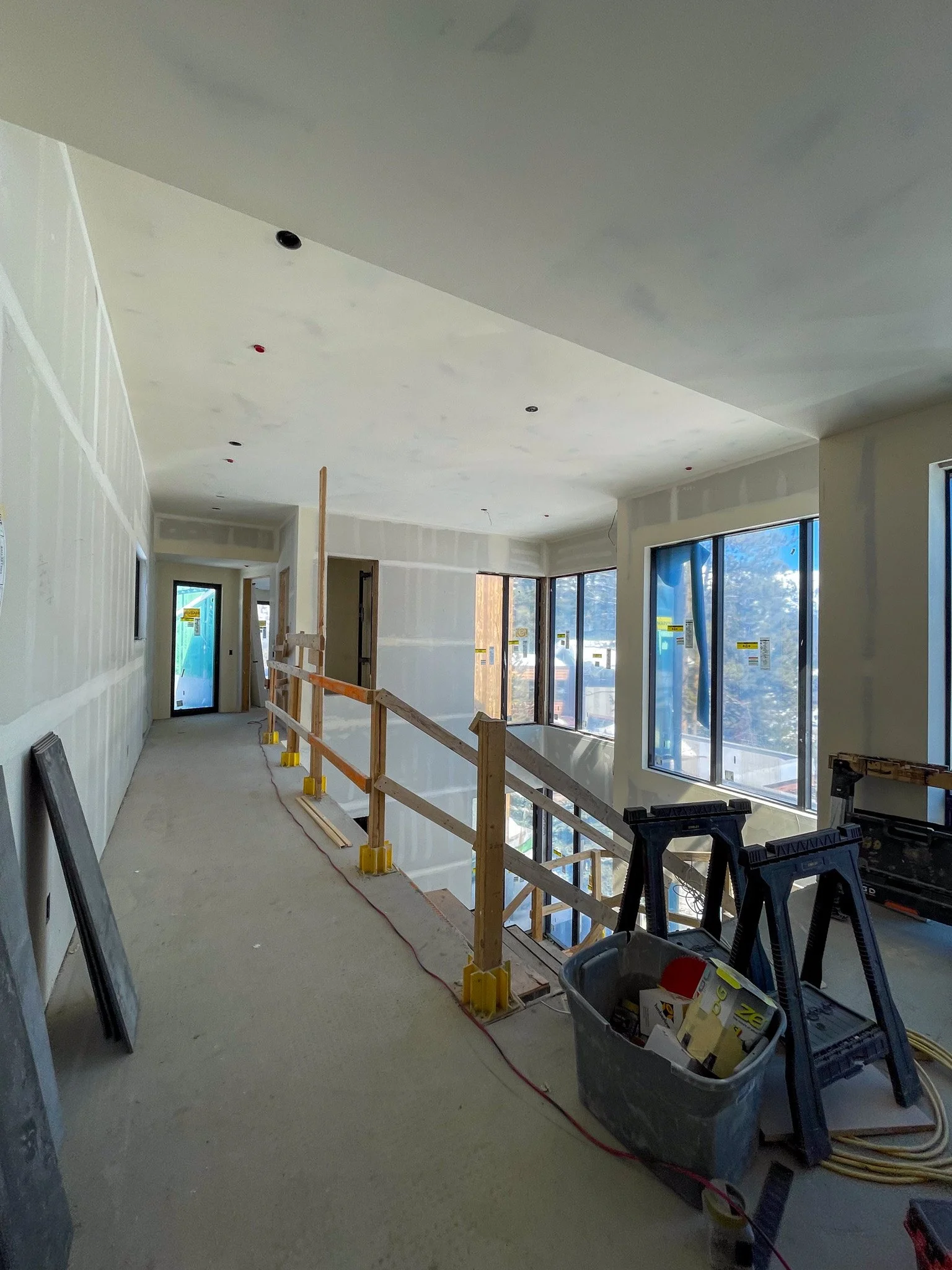 Interior of a building under construction, with drywall walls, large windows, and a temporary wooden railing along a hallway.