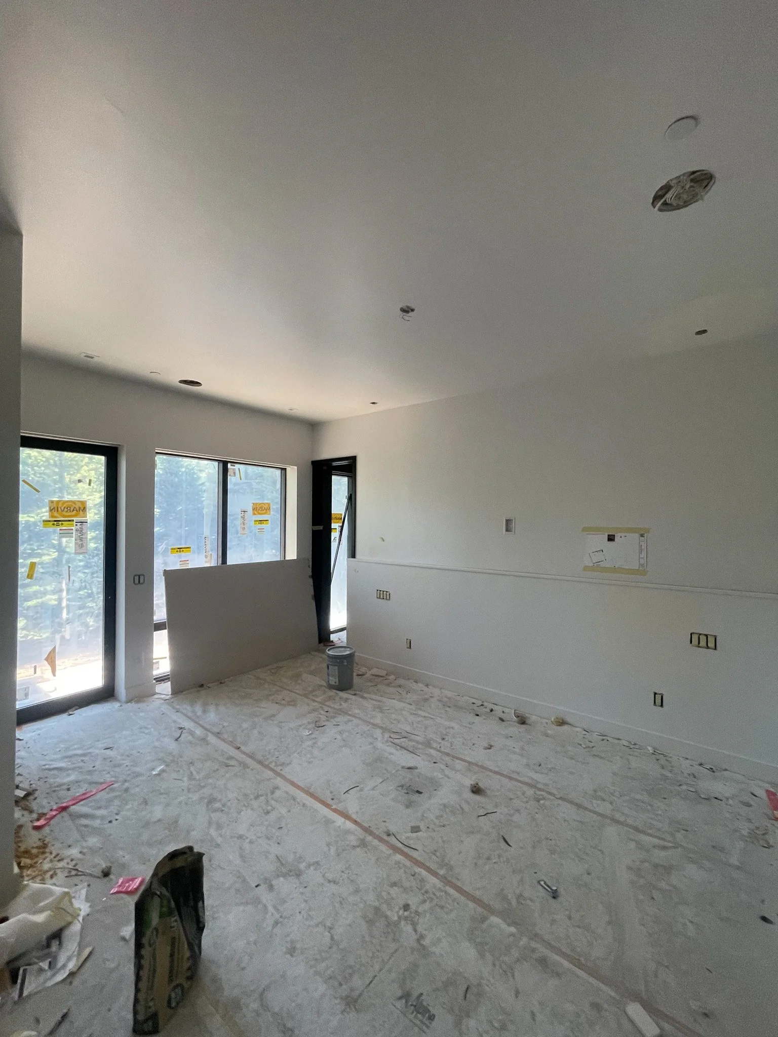 Interior of a room under construction with white walls, large windows, and a concrete floor covered with dust and construction debris.