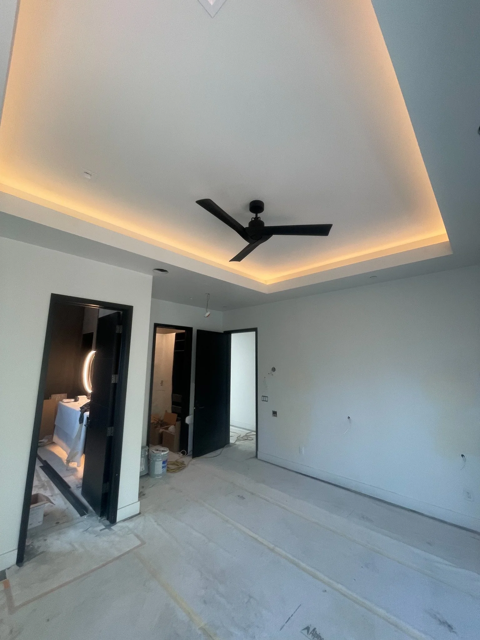 The image shows a modern, unfinished room with a ceiling fan, recessed lighting, and a tray ceiling with LED strip lights. The floor is covered with protective material, and there are open doorways leading to other rooms, with construction supplies p