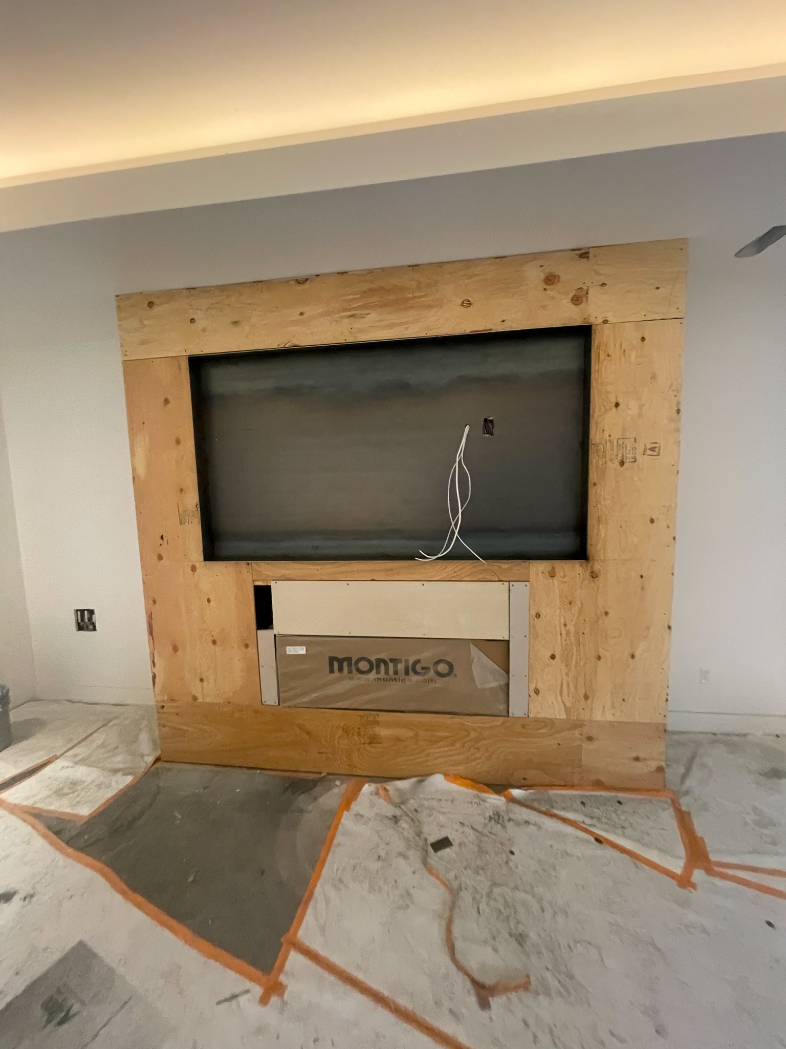 Living room fireplace with a wooden frame under construction, with wires hanging and a protective covering on the fireplace insert, and construction materials on the floor.
