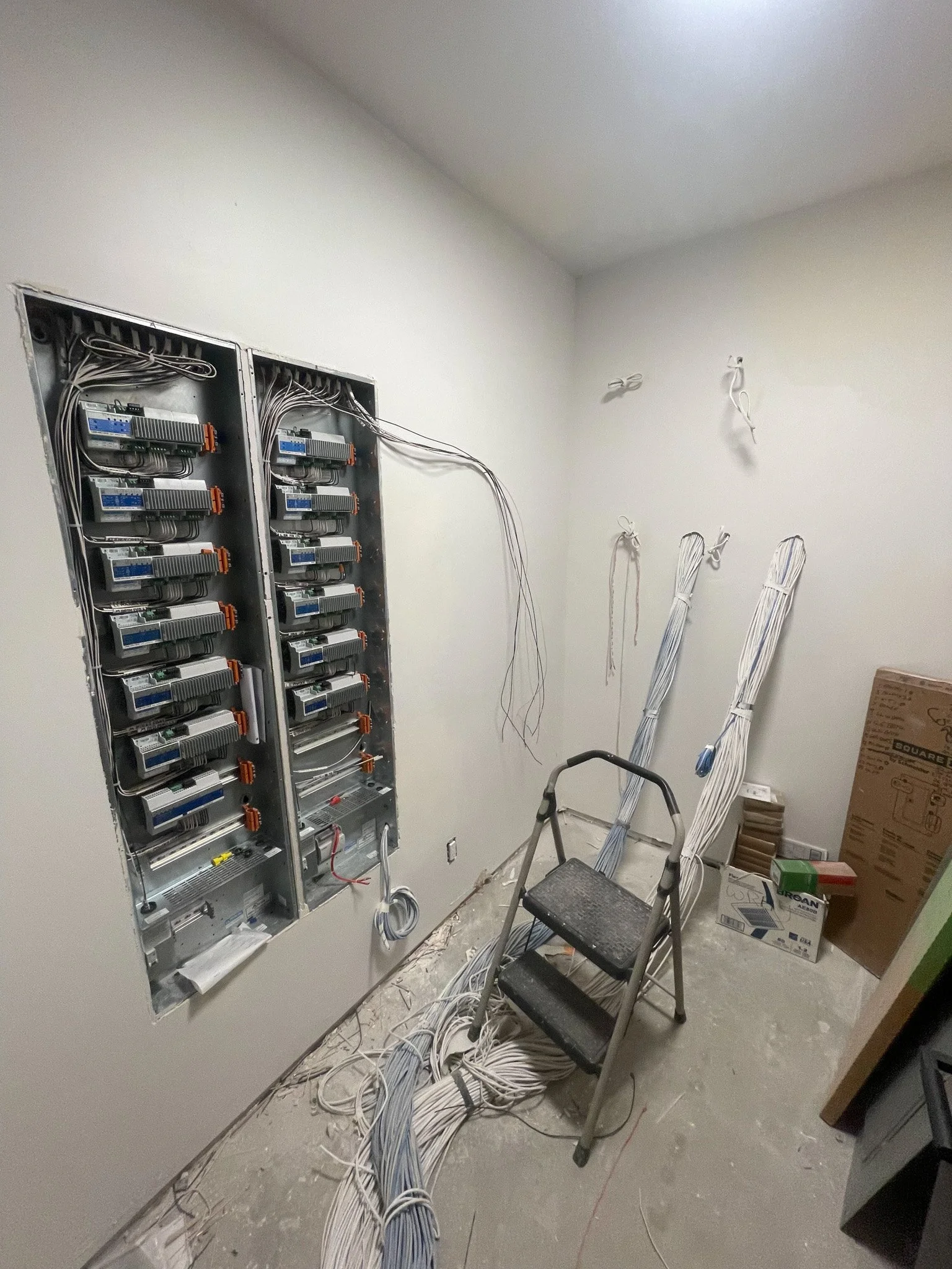Interior of a room under construction with an electrical panel, wires, and tools, including a step ladder and boxes.