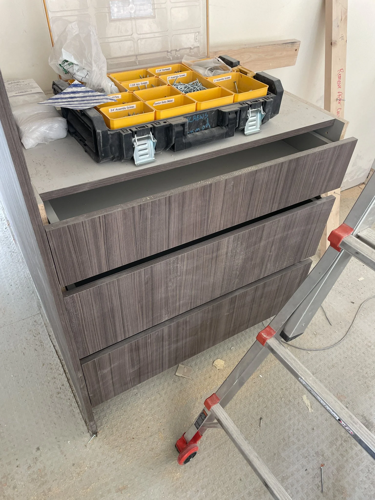 A brown wooden cabinet with three drawers, partially open, situated on a construction site. On top of the cabinet, there is a black plastic toolbox filled with various yellow and clear plastic organizers containing screws, bolts, and other hardware. 