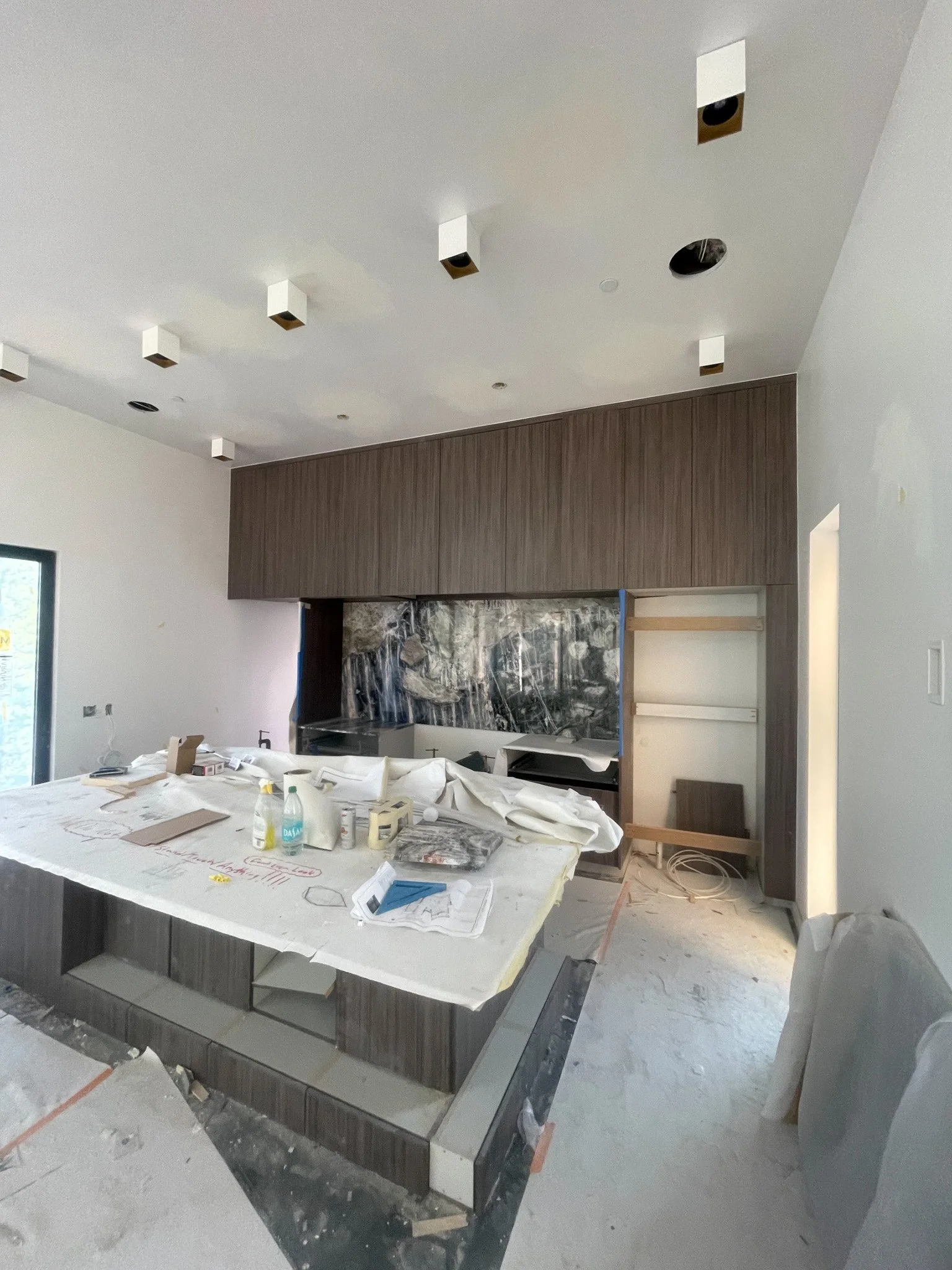 Construction site of a modern living room with a kitchen island covered in white paper, tools, and supplies, and ceiling lights in progress.