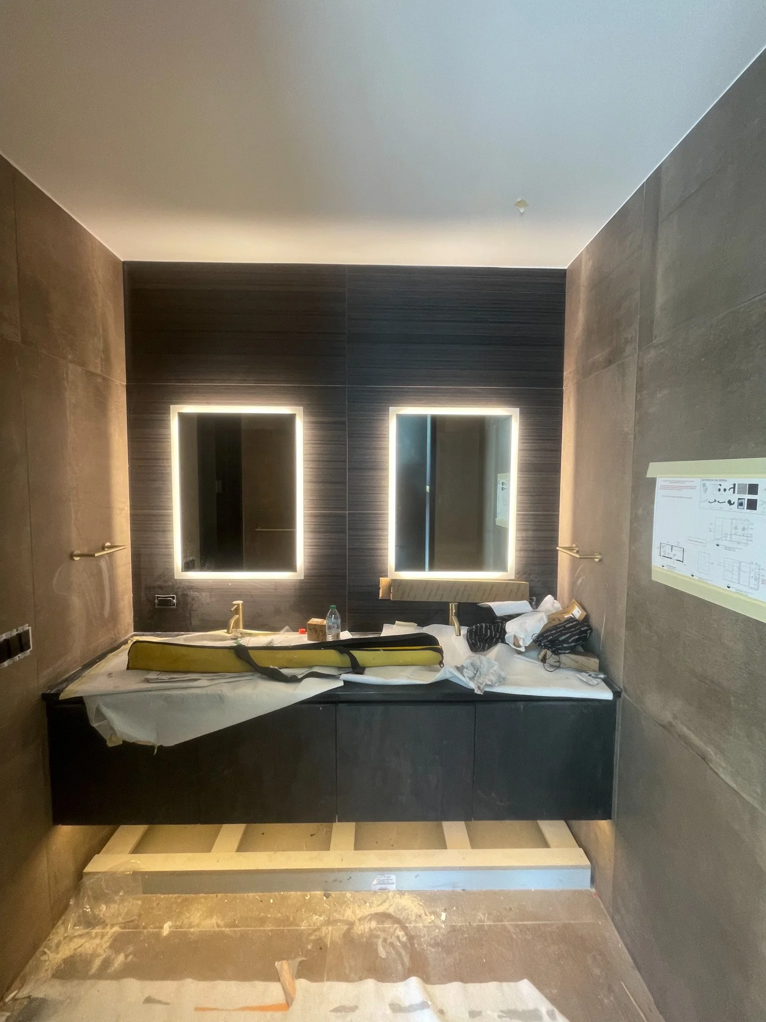 Bathroom vanity with dual illuminated mirrors, dark cabinetry, and ongoing construction.