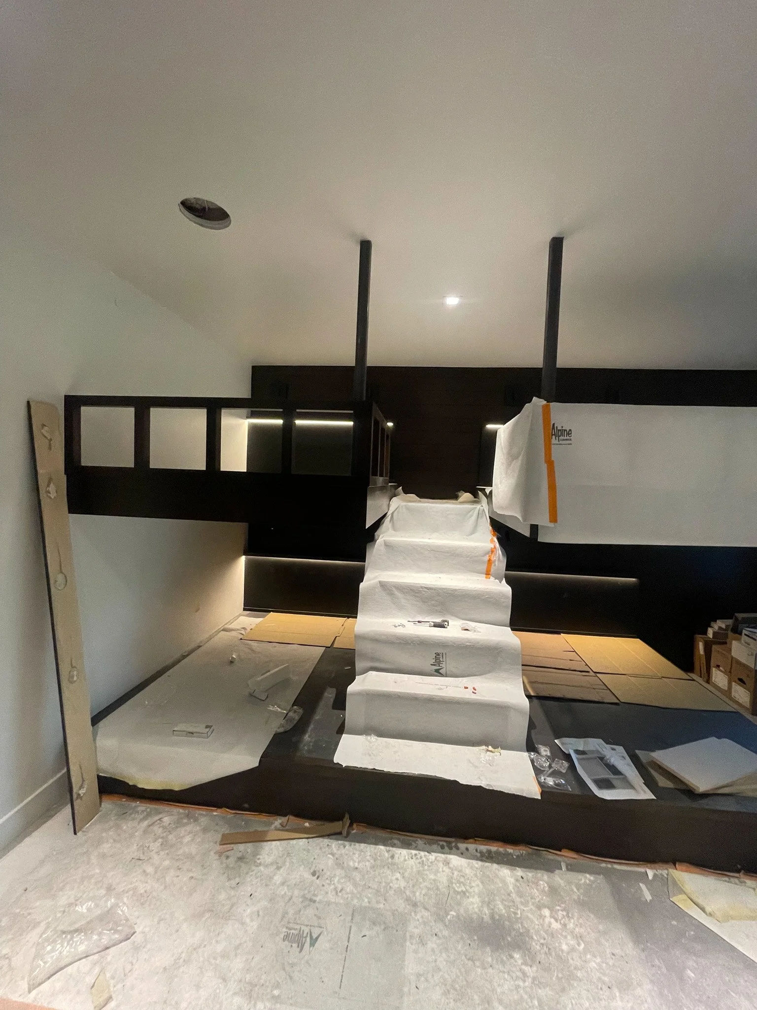 Under construction interior with a staircase covered in protective paper, dark wood paneling, and construction materials scattered on the floor.